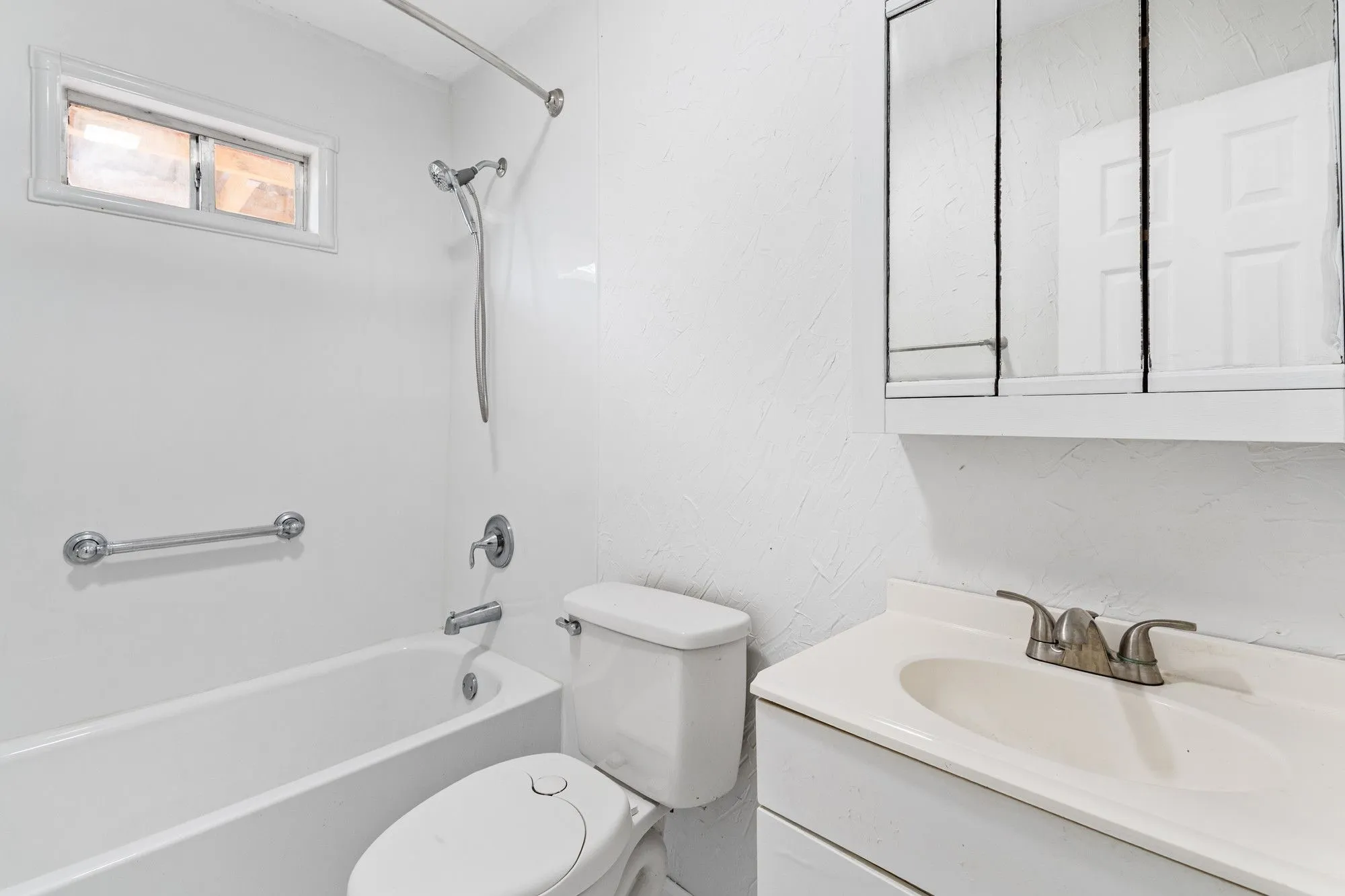 Bathroom with a white tub-shower combination, a window with frosted glass, a wall-mounted grab bar, a white toilet, and a vanity with an integrated sink and mirrored medicine cabinet