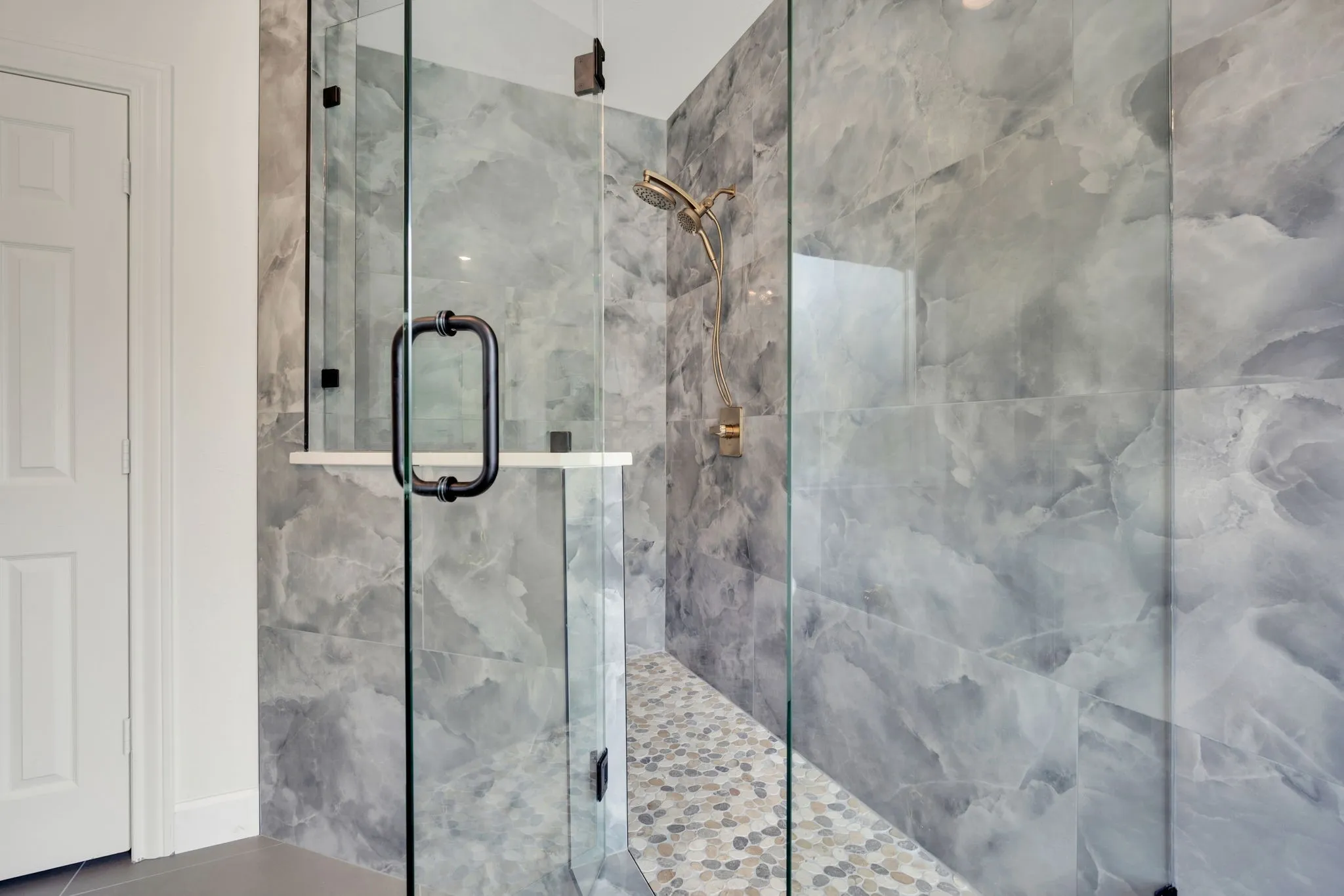 Walk-in shower featuring clear glass enclosure, with black hardware, large-format grey marble-look tile walls, pebble-style floor, and a built-in shower niche