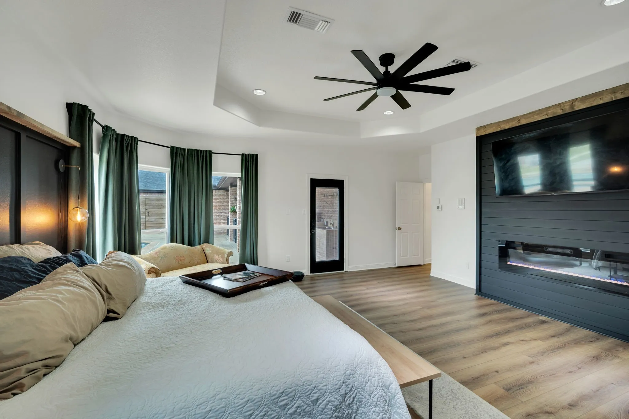 Spacious primary room featuring wood-finish flooring, a contemporary ceiling fan, electric fireplace, and a tray ceiling