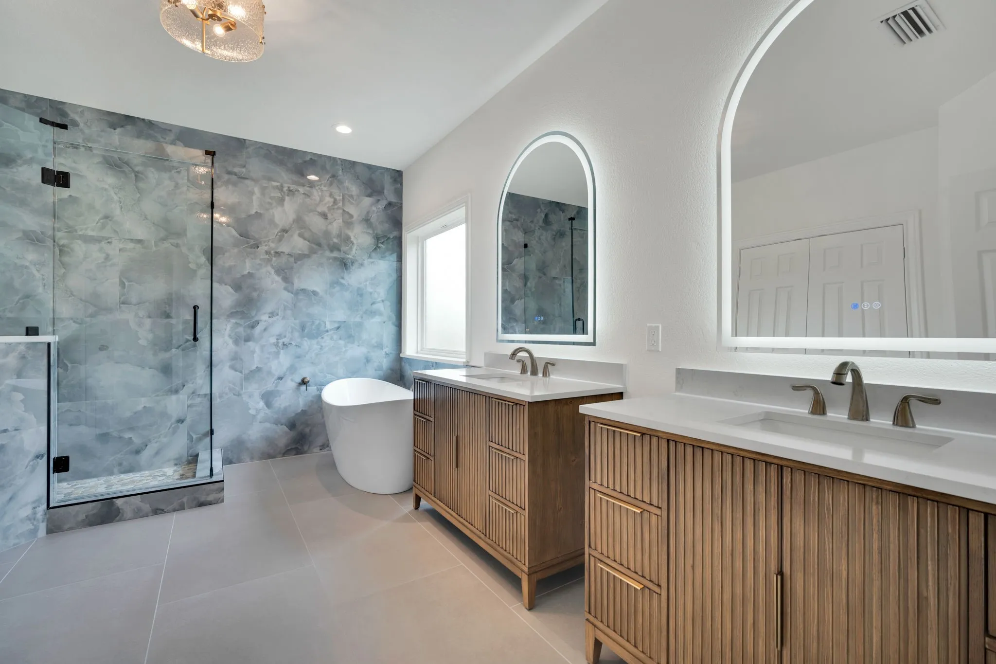 Modern bathroom featuring a freestanding soaking tub, a spacious glass-enclosed shower with a beautiful tile wall, dual wood-finish vanities with countertop sinks, and illuminated arched mirrors