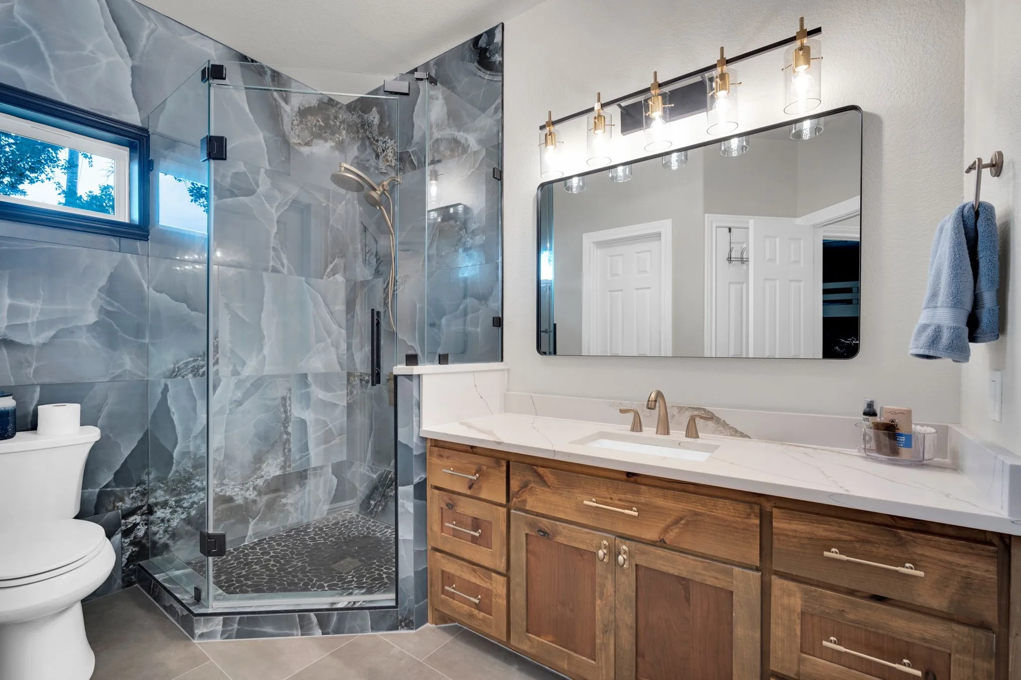 Bathroom 5 featuring a glass-enclosed shower with large format stone-finish wall tiles and a pebble floor, a wood-finish vanity with a quartz countertop and undermount sink, and a rectangular mirror with a five-light fixture