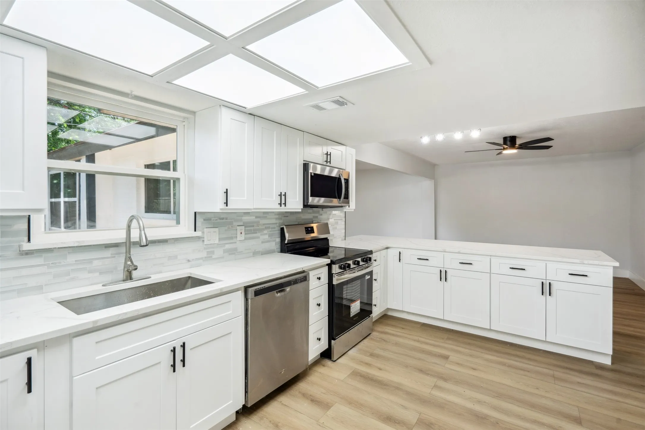Contemporary kitchen featuring white shaker cabinetry, stainless steel appliances, light-toned wood-finish flooring, a tile backsplash, and white countertops