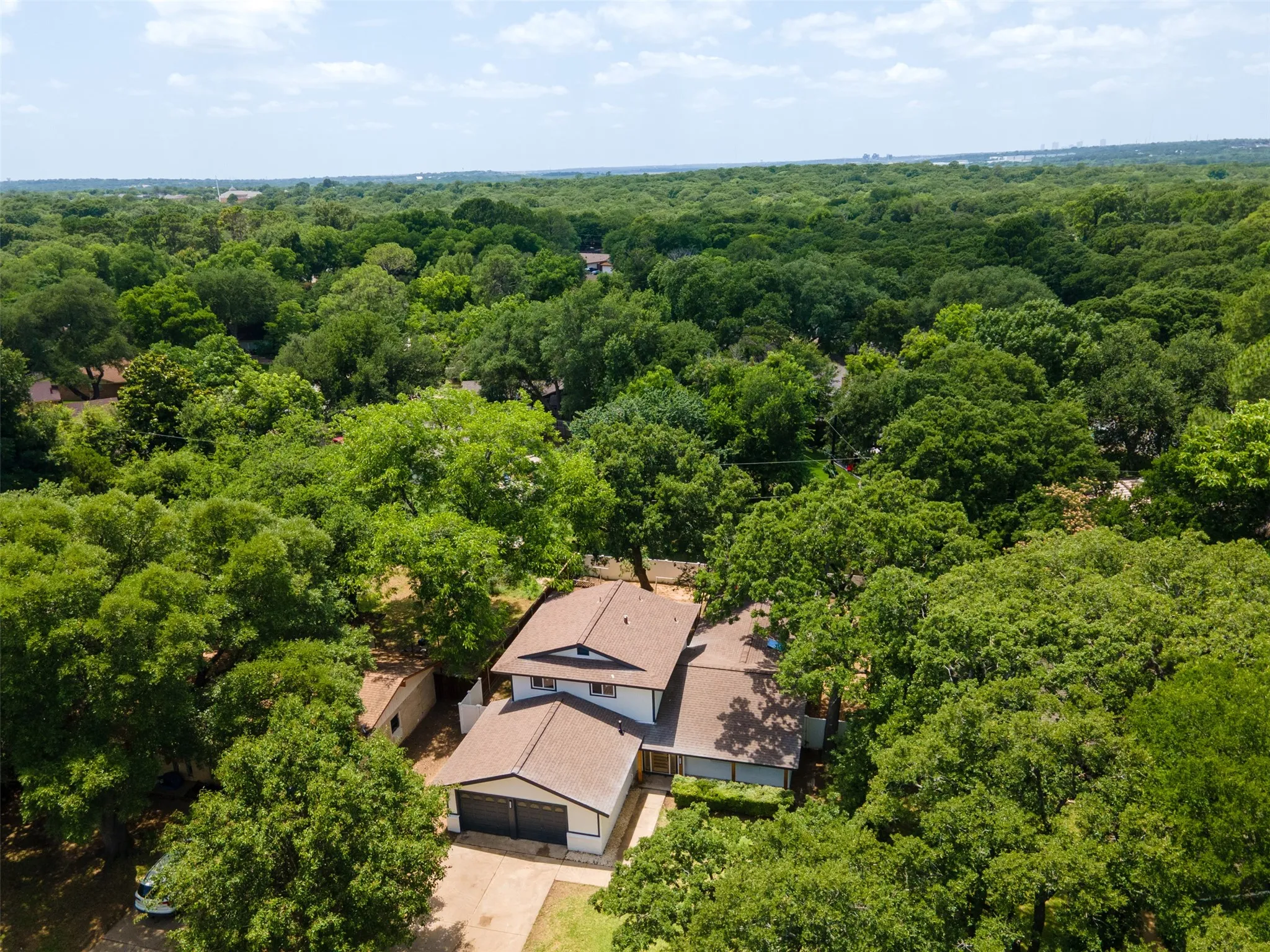 Expansive tree canopy surrounding a property featuring a multi-level structure with a brown shingled roof, white siding, and a two-car garage