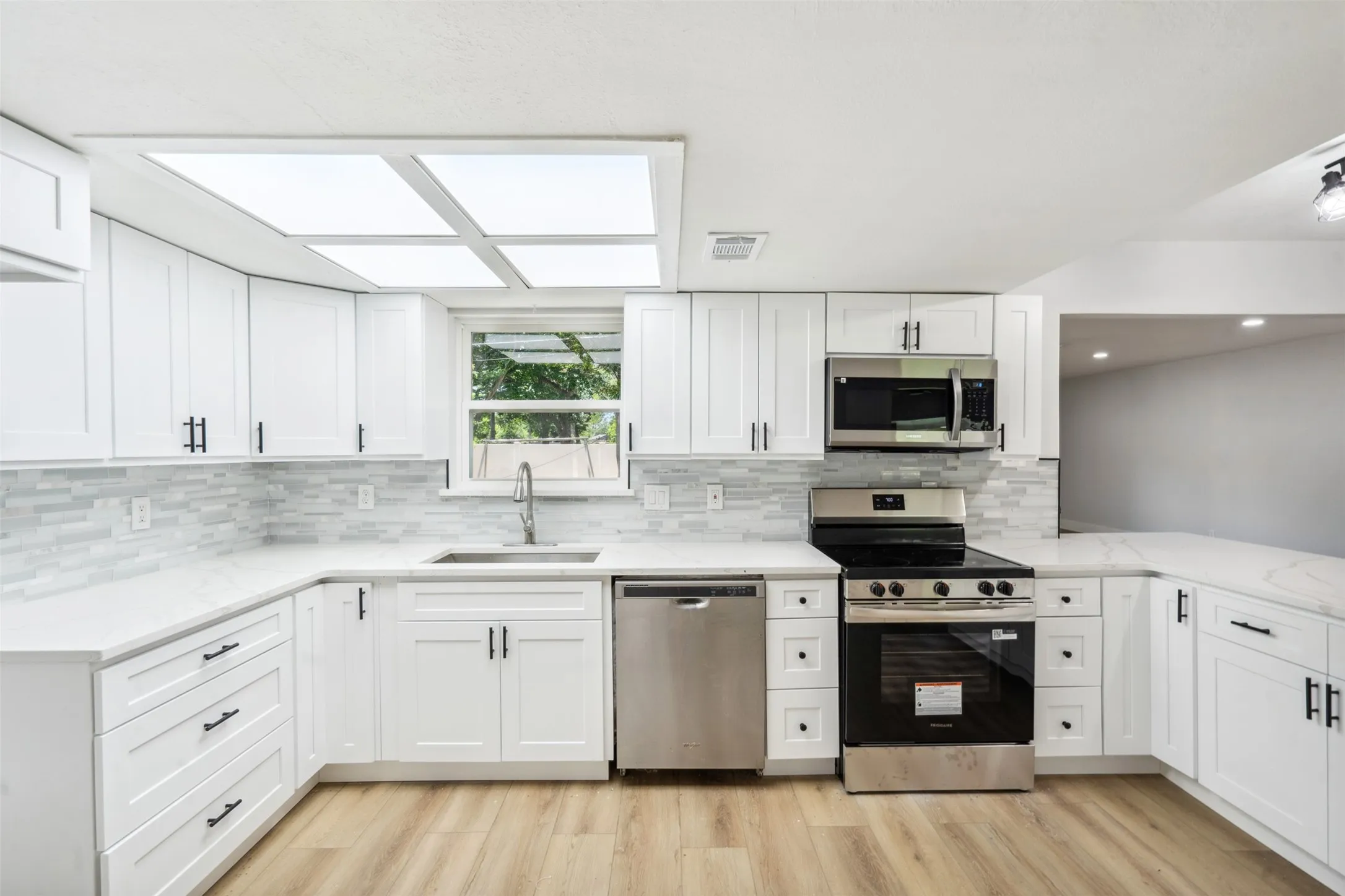 Contemporary kitchen featuring white shaker cabinetry, stainless steel appliances, light-toned wood-finish flooring, white countertops, and a subway tile backsplash