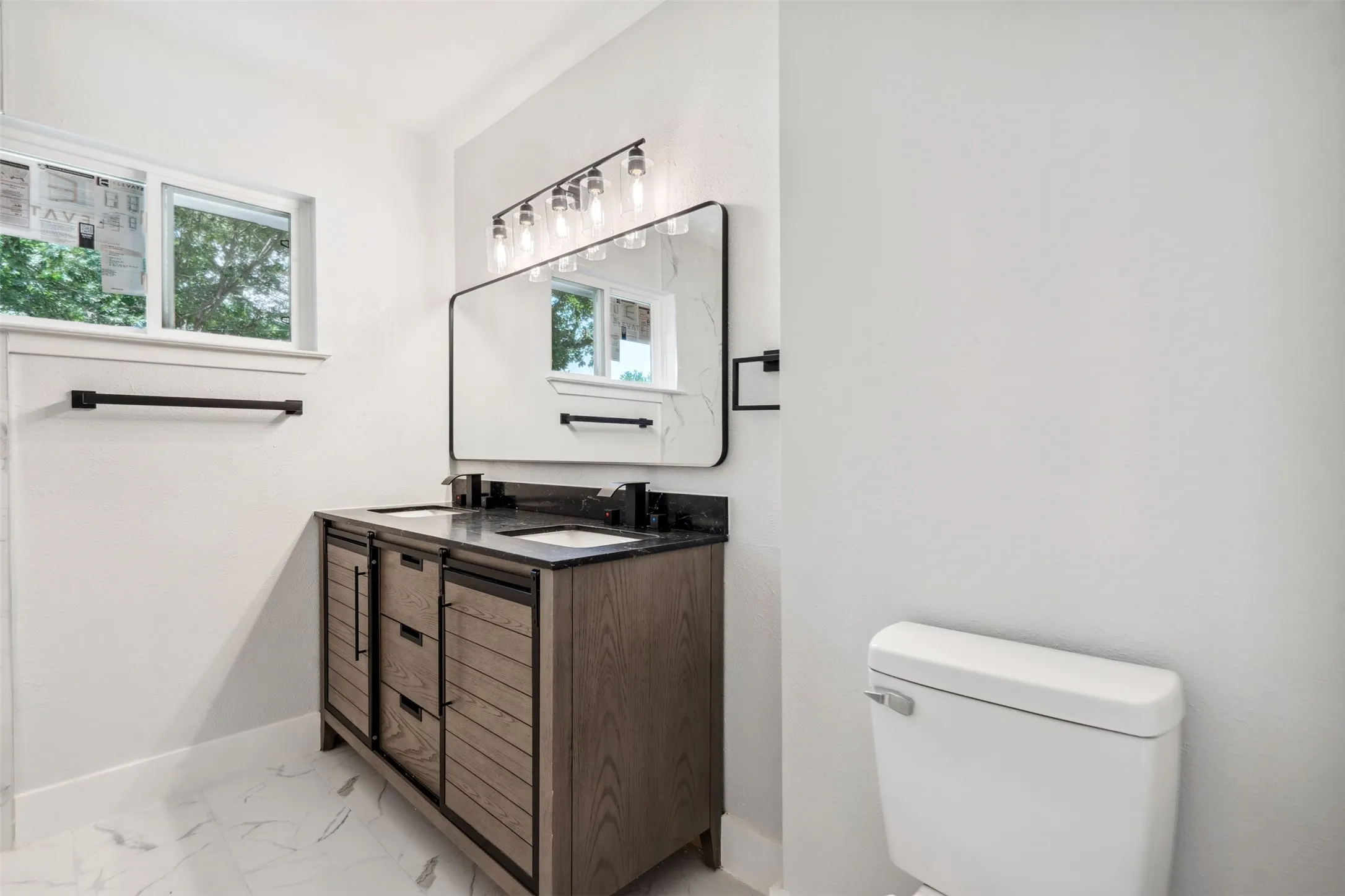 Bathroom featuring a dual-sink vanity with a dark countertop and wood-finish cabinetry, a large framed mirror, and a multi-bulb vanity light fixture
