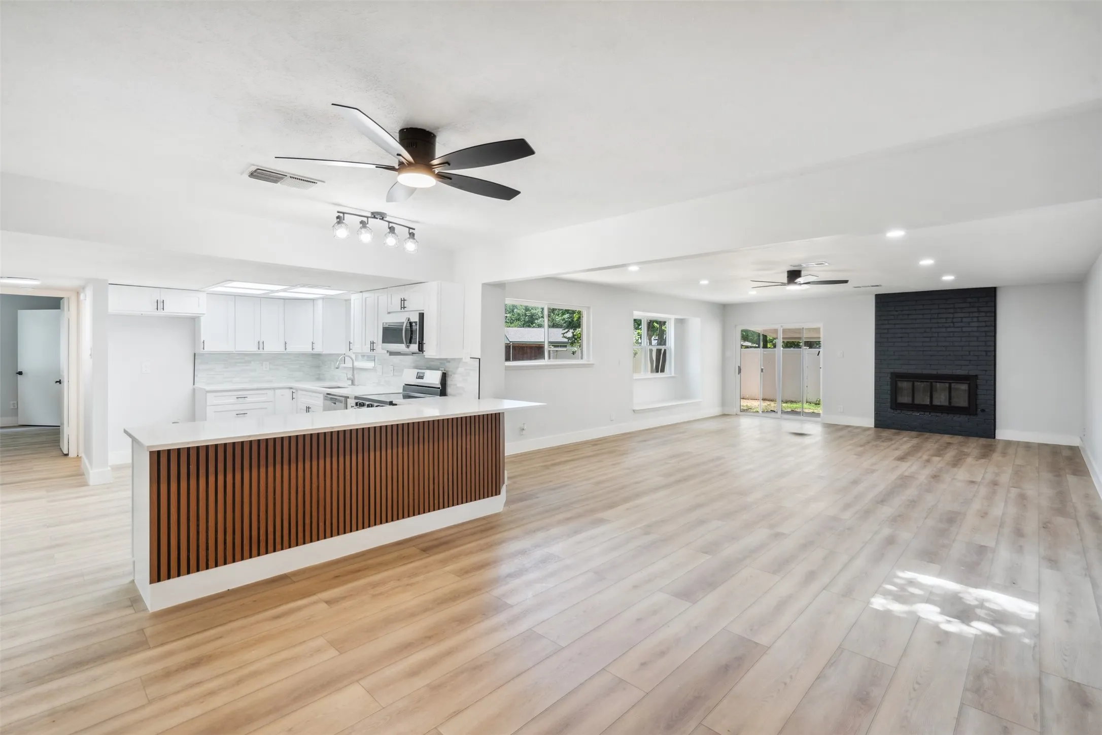 Open-concept living area featuring wood-finish flooring throughout, a modern kitchen island with slatted wood-look paneling and a light-toned countertop, white cabinetry with stainless steel appliances, and a black brick fireplace with a glass enclosure