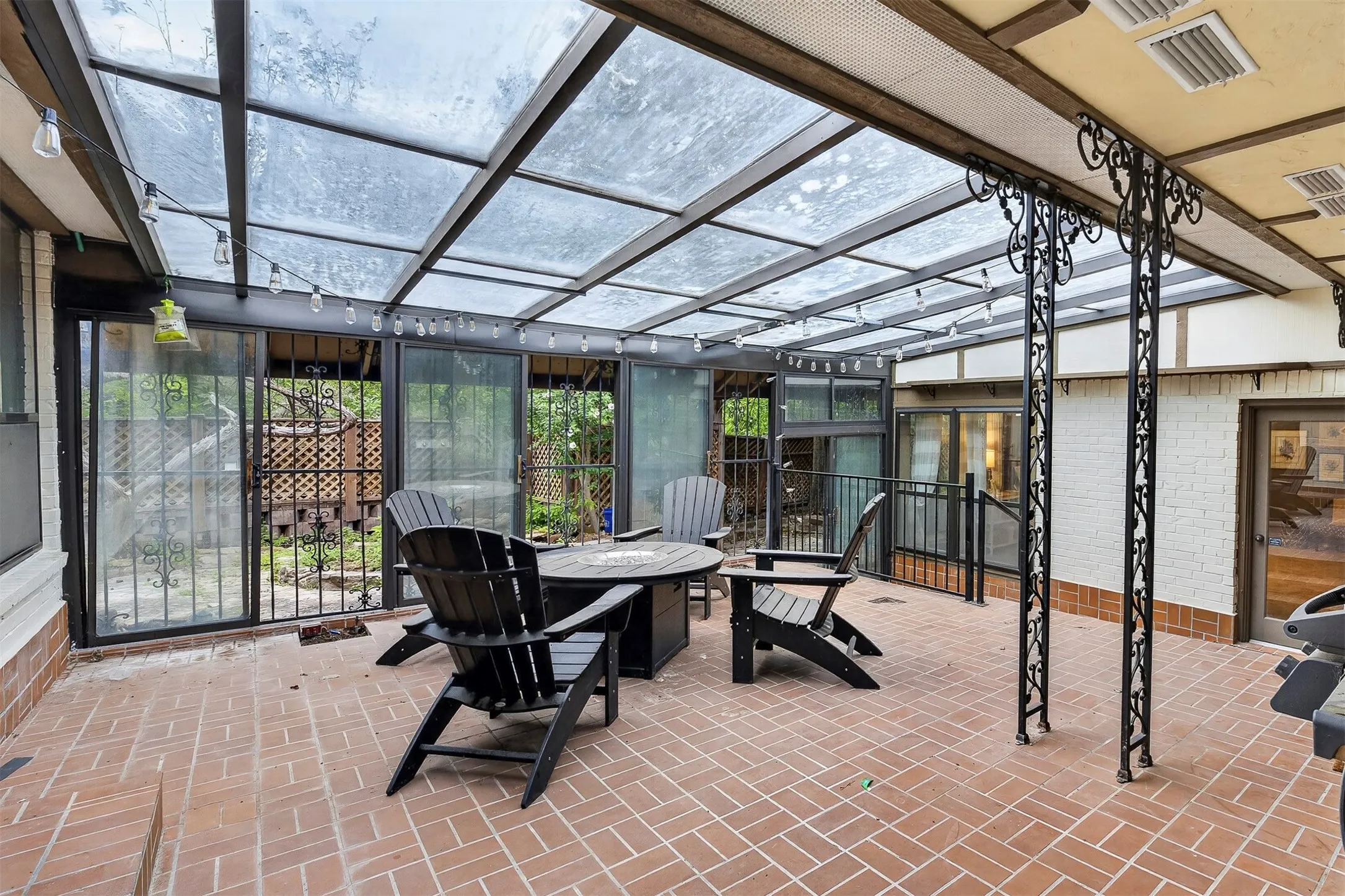 Enclosed patio featuring a clear panel roof, brick flooring in a herringbone pattern, sliding glass doors with security grates, and decorative wrought iron pillars