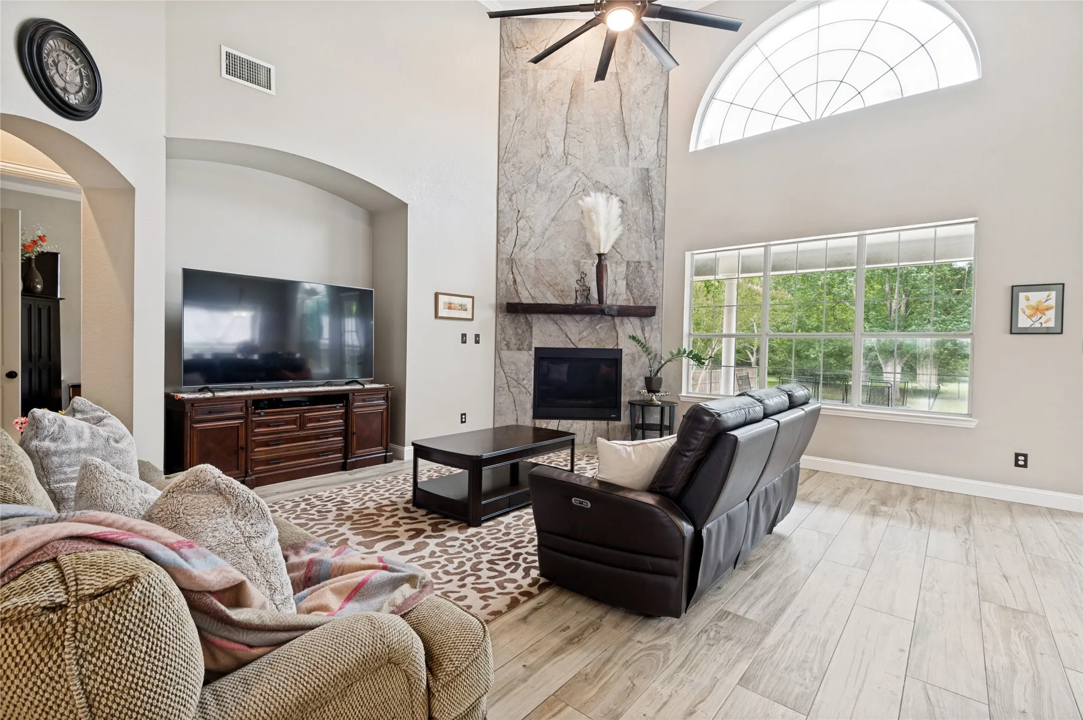 Vaulted living room featuring a stone fireplace with a wood mantel, a ceiling fan, and large windows with an arched transom