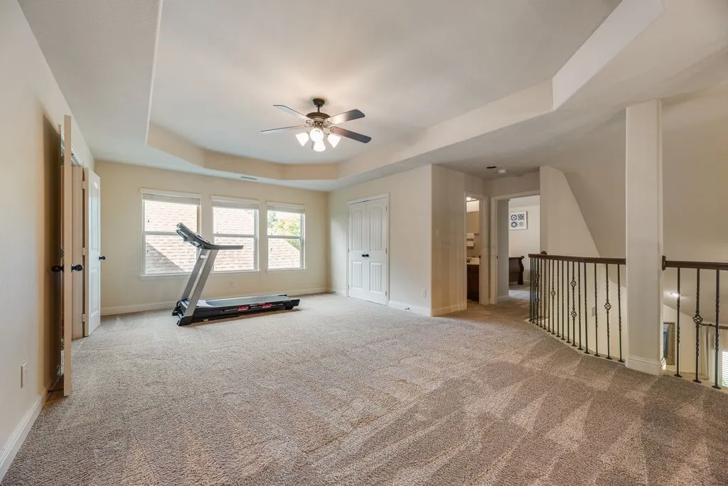 Spacious carpeted room featuring a tray ceiling with a ceiling fan, multiple windows, and a wrought iron banister overlooking a lower level
