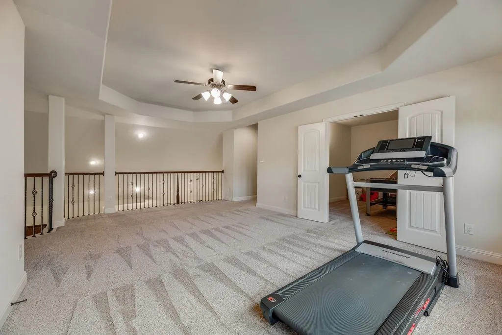 Spacious carpeted room featuring a tray ceiling with a ceiling fan, multiple windows, and a wrought iron banister overlooking a lower level