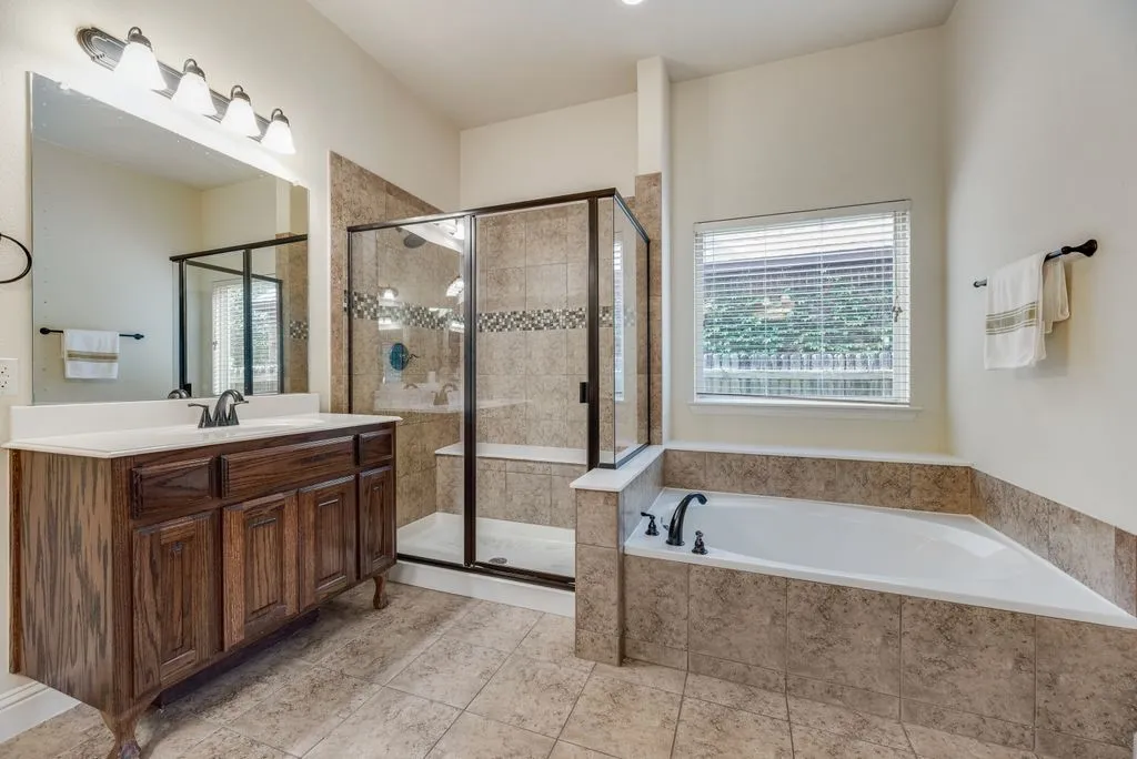 Bathroom featuring a wood-finish vanity with a single basin, a large mirror, a tiled walk-in shower with glass enclosure, a built-in tub with tile surround, and a window with blinds