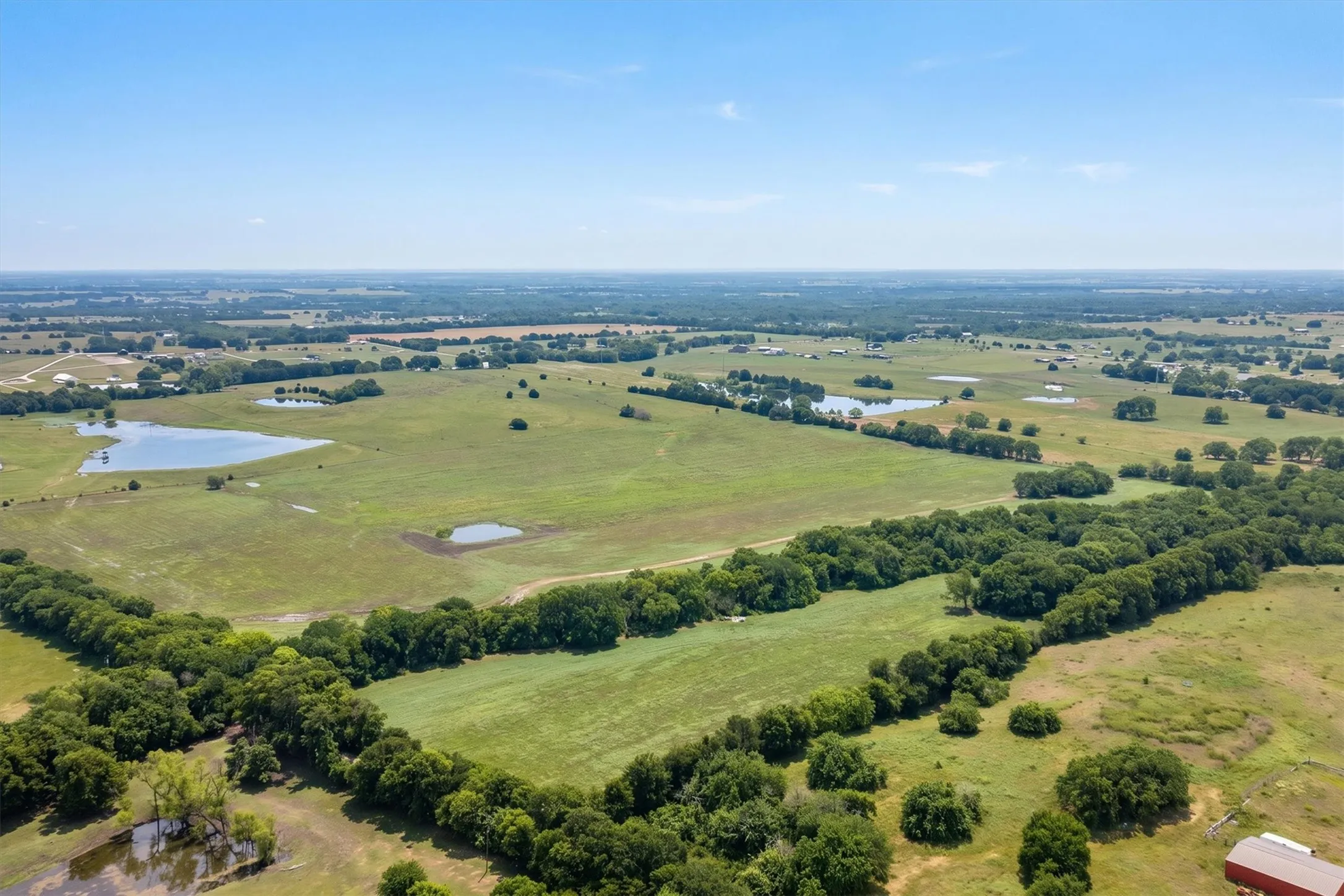 Expansive rural landscape featuring extensive green pastures, multiple natural ponds, scattered mature trees, and a distant red barn structure