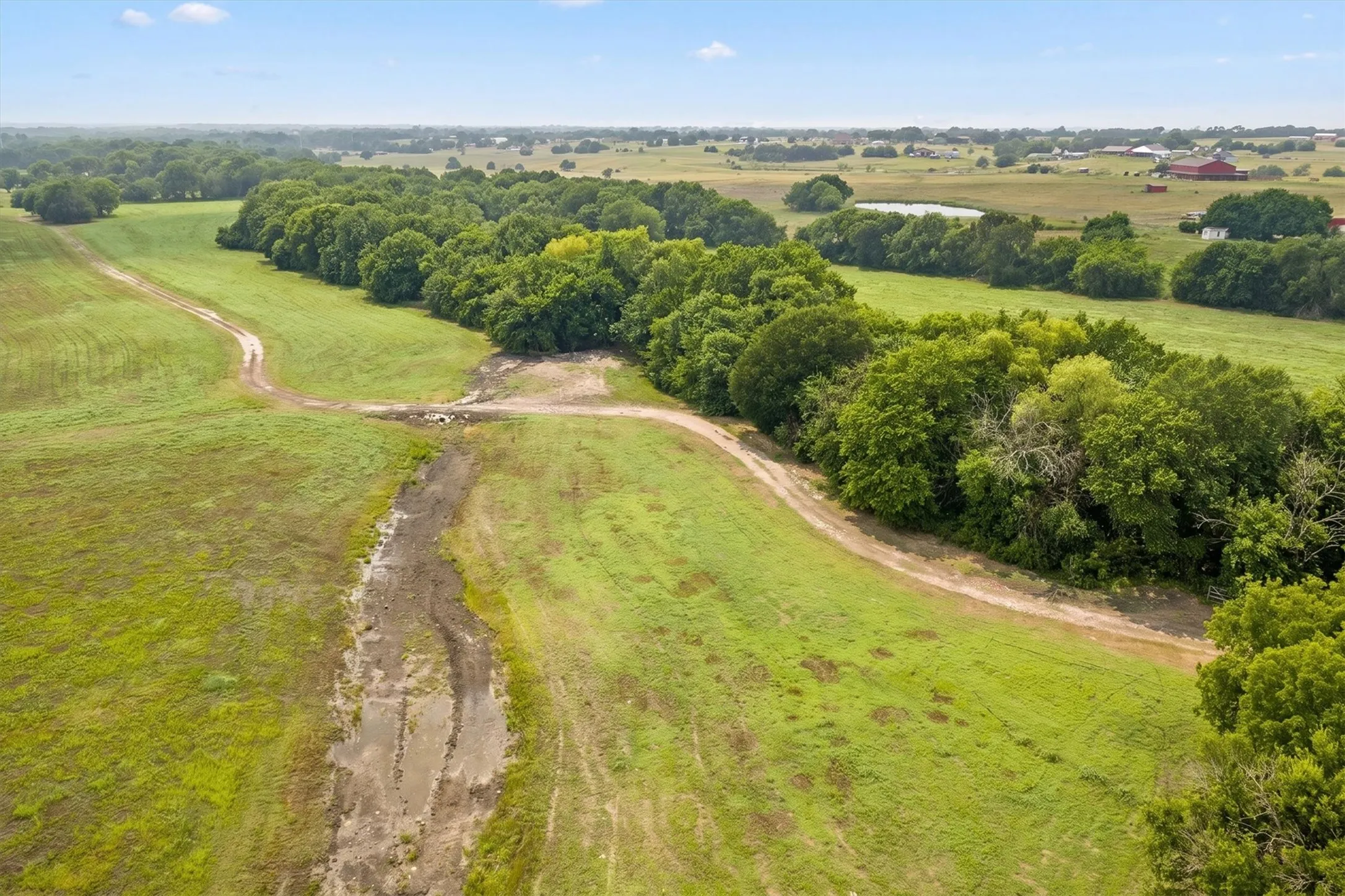 Expansive green acreage featuring a meandering dirt path, mature tree lines, and distant agricultural structures