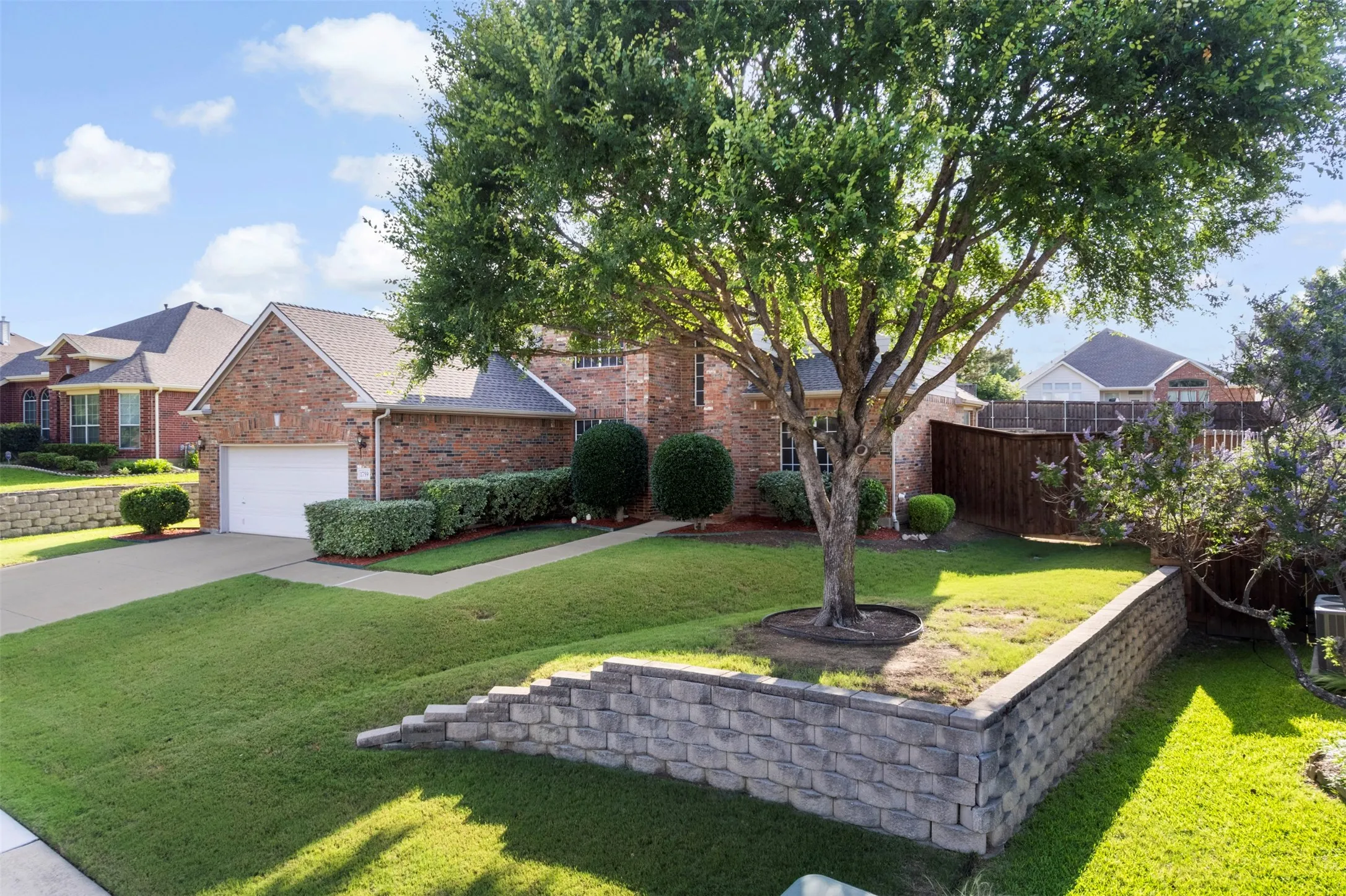 Brick exterior residence featuring a front-facing garage with a white door, mature tree, manicured lawn, and stone block retaining walls