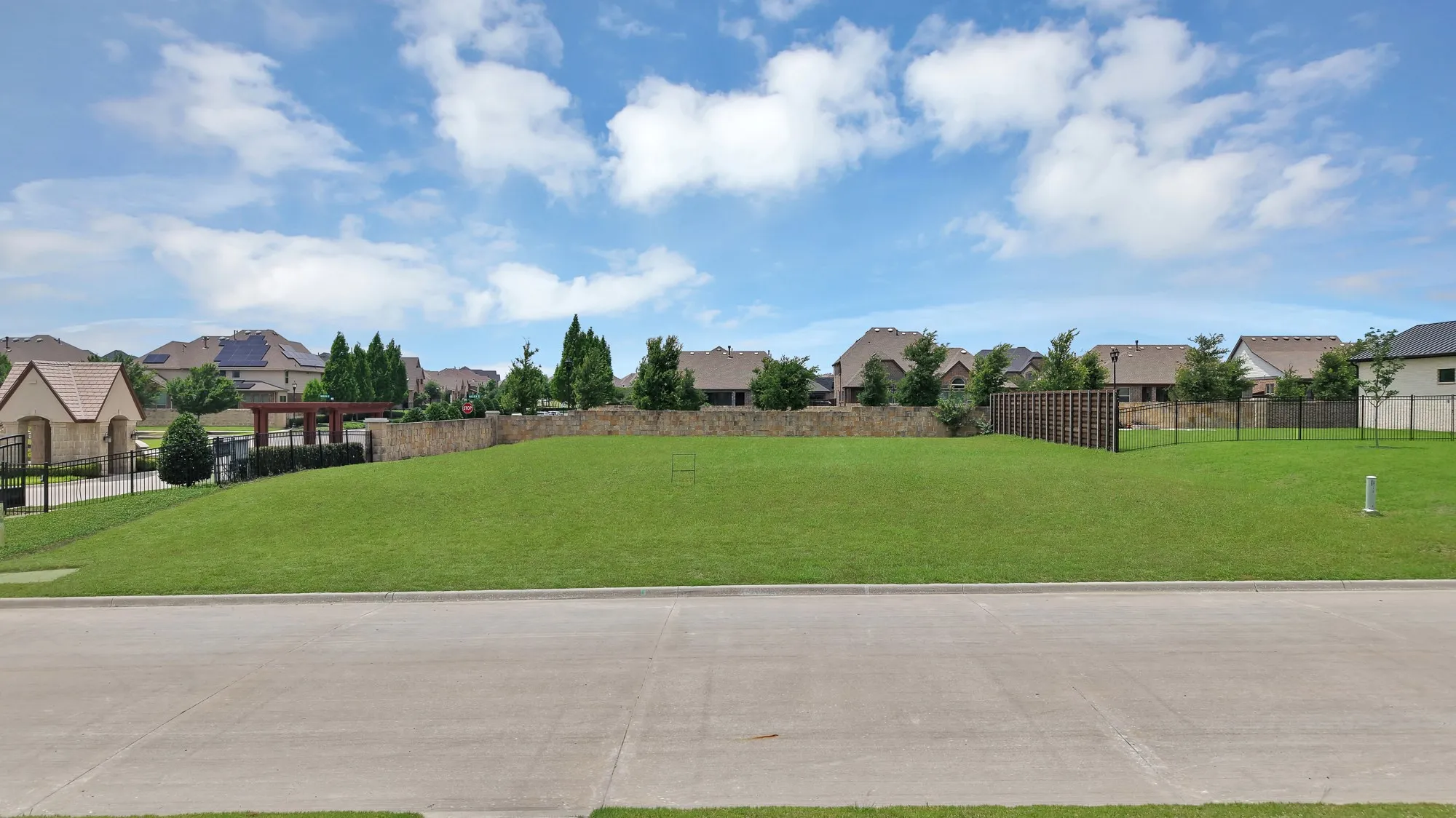 Lot street view — Level, sodded lot with full concrete curb and sidewalk. Stone perimeter community wall along rear, iron fencing on adjacent parcel, and cedar pergola visible at neighboring gate structure.
