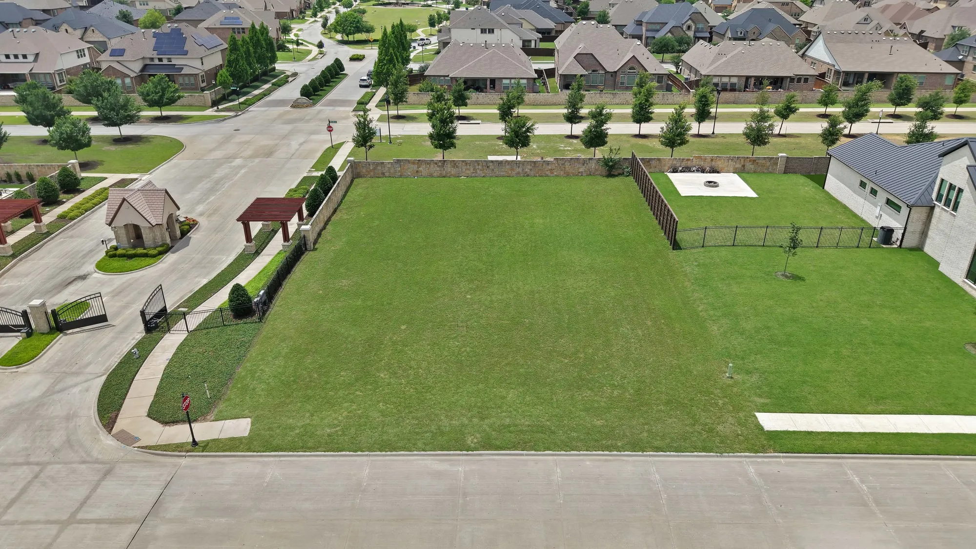 Lot aerial with fire pit pad — Nearly half-acre lot, cleared and sodded, with concrete fire pit pad visible on adjacent home's property for scale reference. Gated entry kiosk and pergola structure directly to the left. Curb cut and sidewalk stub in place.