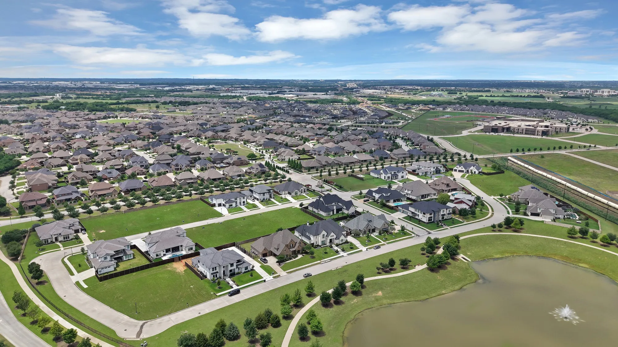 Neighborhood aerial with pond — Expansive aerial view of South Pointe showcasing mix of custom home styles, tree-lined streets, community pond with fountain, and open green corridors. Active development growth visible to the east.