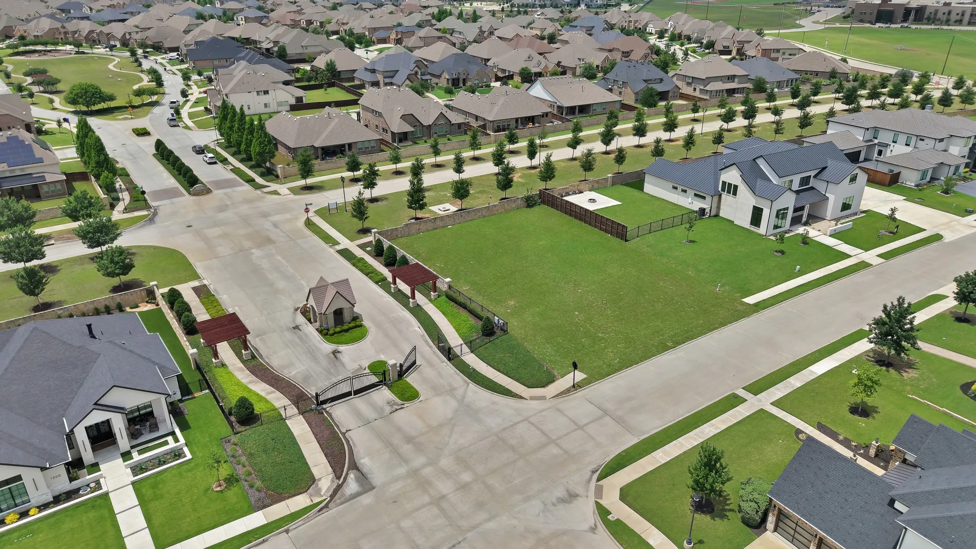 Lot and gated entry aerial — Lot positioned directly across from the South Pointe gated entrance with pergola and guard house. Stone perimeter wall along rear boundary, iron fencing along the adjacent neighbor's property line, and open street frontage.