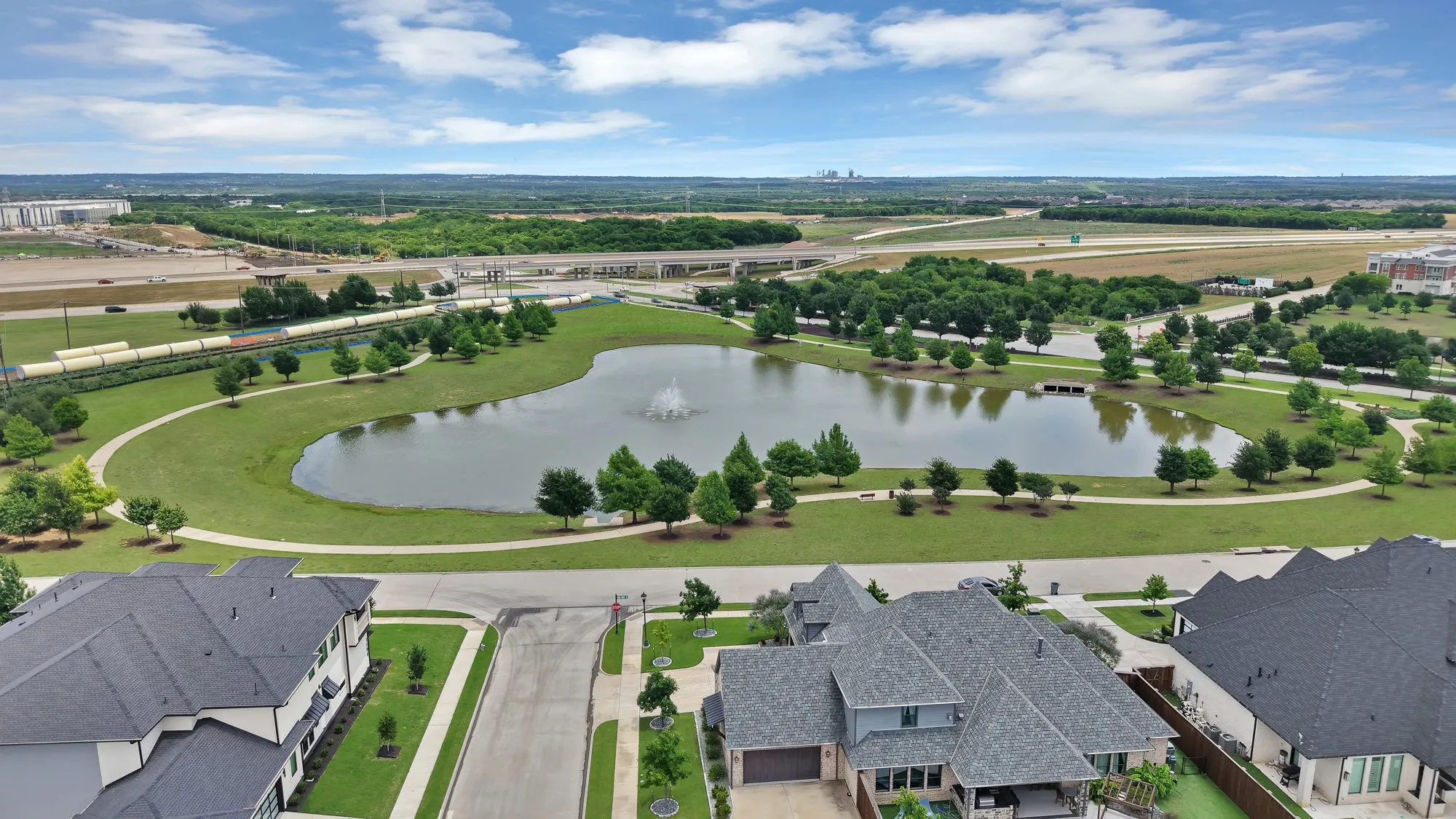 Community pond and park — Stocked retention pond with aerating fountain feature surrounded by mature tree-lined walking paths and maintained park greenspace. Interstate visibility and open skyline views beyond.