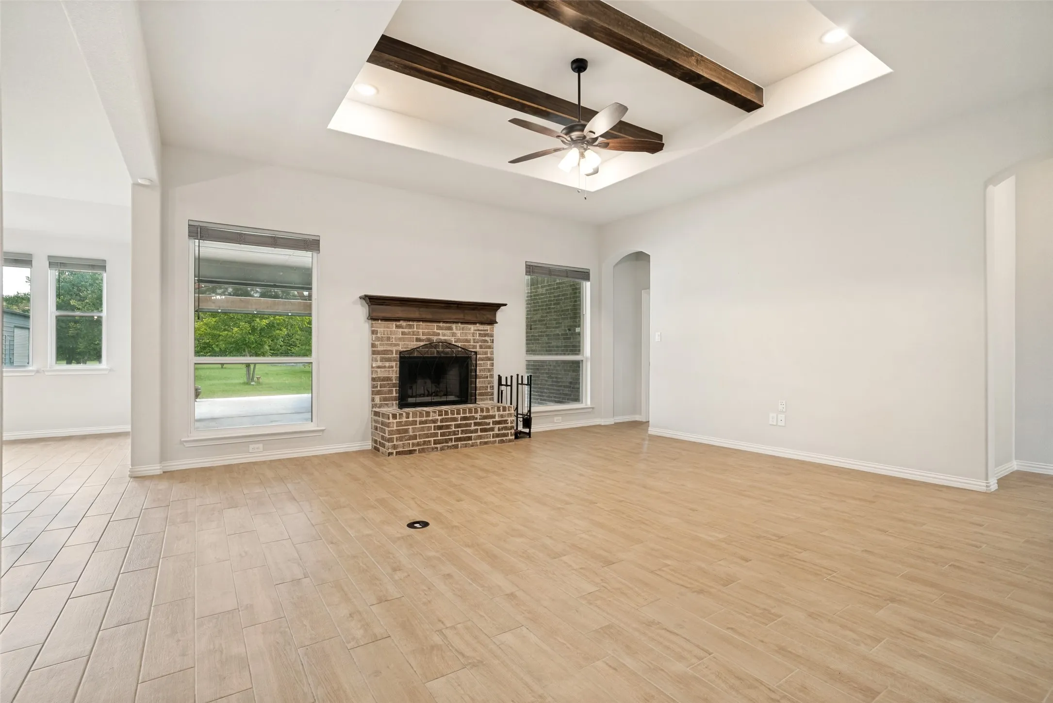 Spacious interior featuring wood-finish flooring, a brick fireplace with a wood mantle, and a tray ceiling with exposed wood beams and a ceiling fan