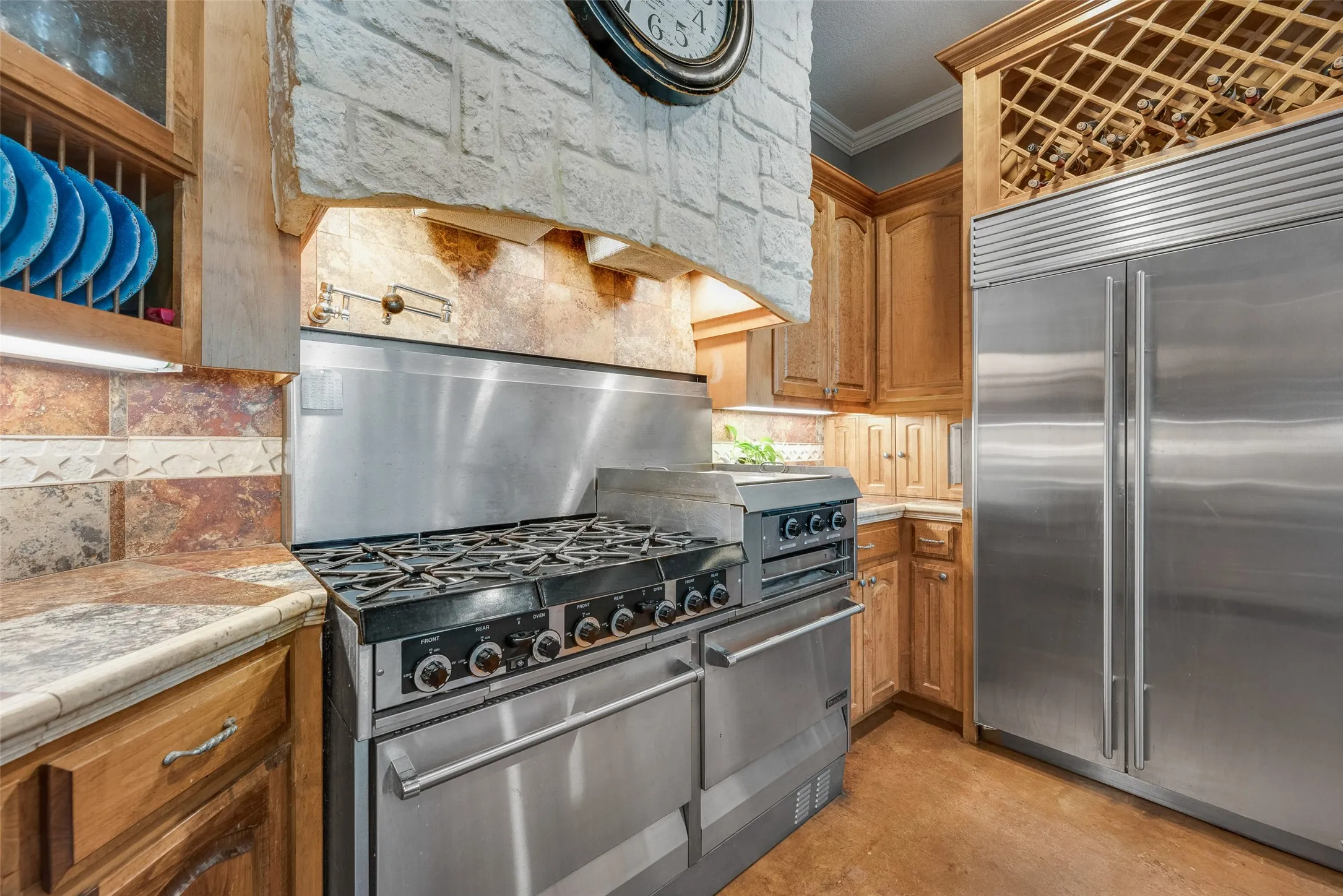 Professional-grade stainless steel range with a built-in griddle, stone-clad range hood, custom wood cabinetry, stainless steel refrigerator, and an integrated wine rack