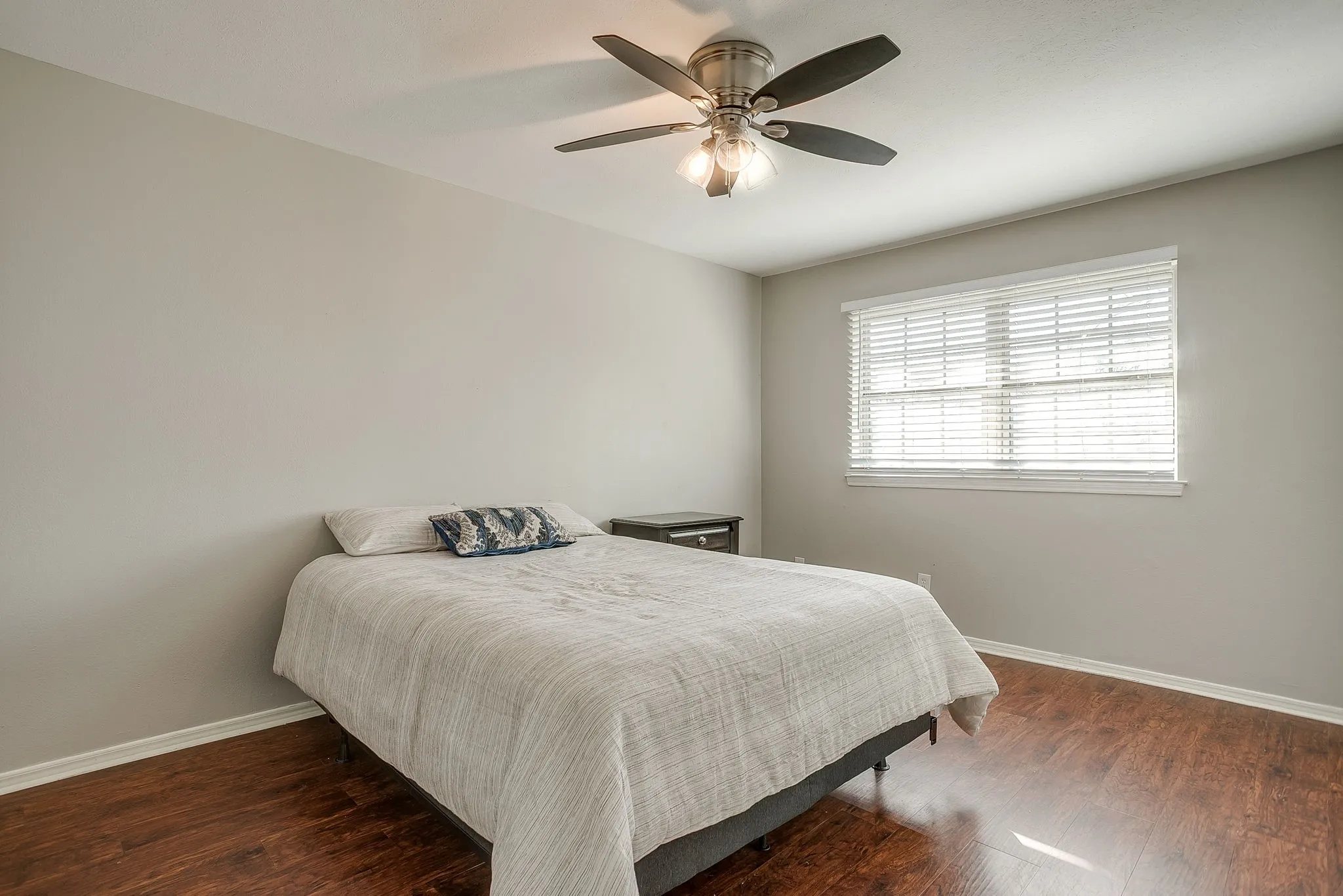 Spacious room featuring wood-finish flooring, a ceiling fan with integrated lighting, and a large window with horizontal blinds