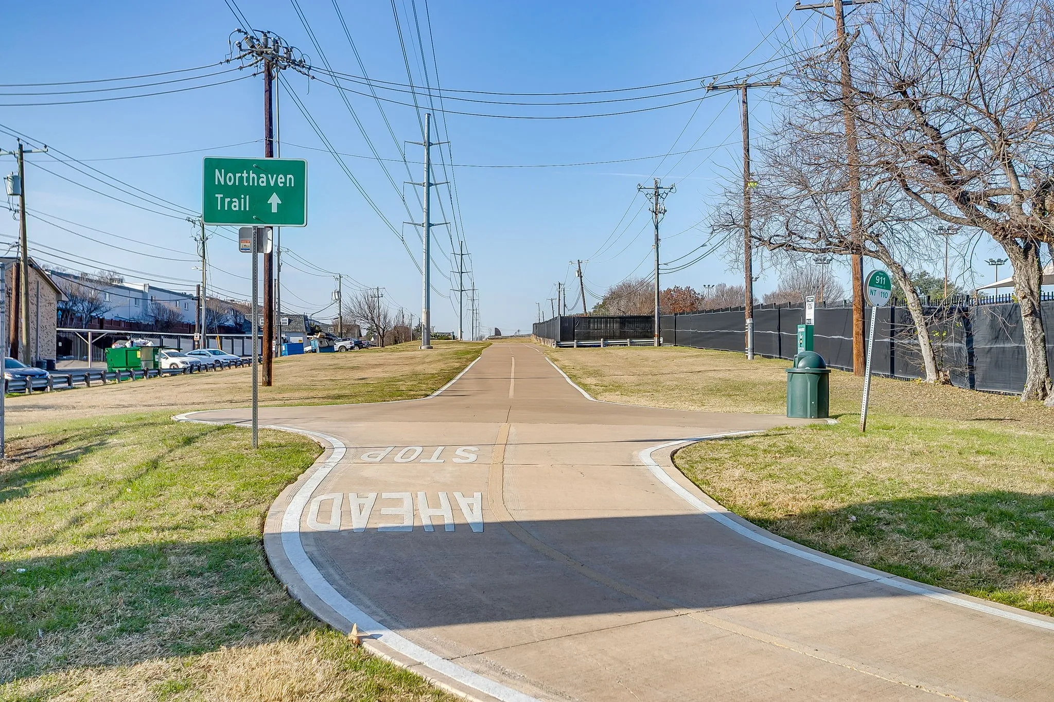 Paved trail with marked intersection and signage, bordered by grass and fencing