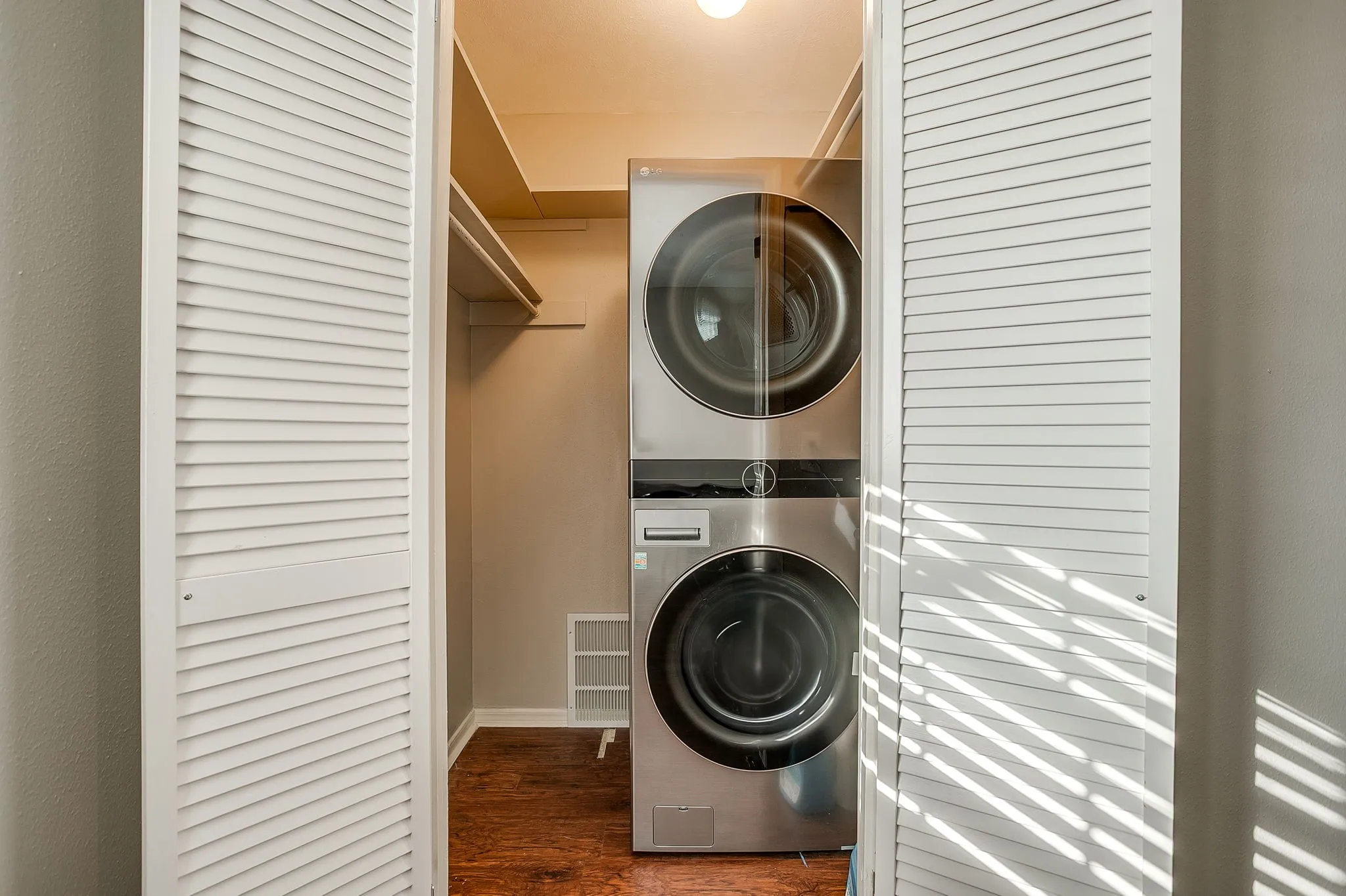 Laundry closet featuring stacked front-loading washer and dryer units, white louvered bifold doors, wood-finish flooring, and built-in shelving