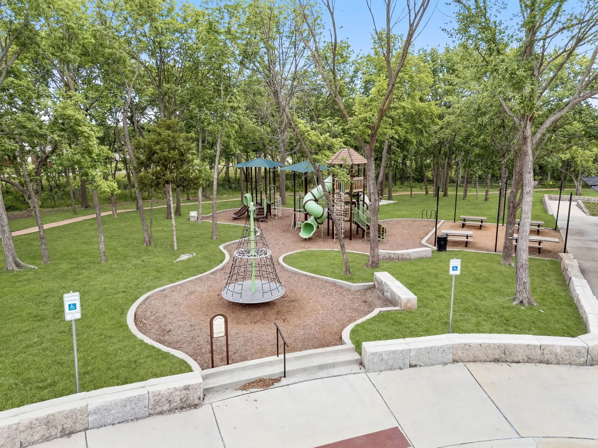 Detailed view of a vibrant community playground area, featuring an assortment of engaging play structures designed for children of various ages, a challenging climbing net for adventurous kids, strategically placed picnic tables perfect for family gatherings or casual outdoor meals, and an expansive, inviting green space offering ample room for free play and outdoor activities, all contributing to a welcoming and stimulating environment for the community.