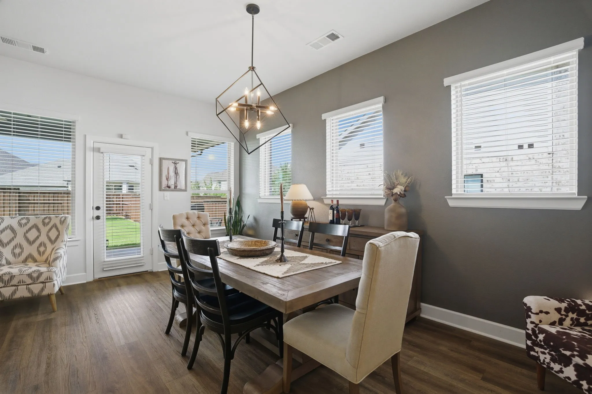 This photo showcases a stylish dining area featuring warm wood-look flooring, complemented by a modern and eye-catching contemporary chandelier that provides ample illumination. The space is flooded with natural light thanks to multiple windows, each equipped with functional and elegant blinds for privacy and light control. A charming glass-paned exterior door adds a touch of sophistication and allows for easy access, while the two-tone wall treatment creates an interesting visual texture and adds a layer of depth and character to the room.