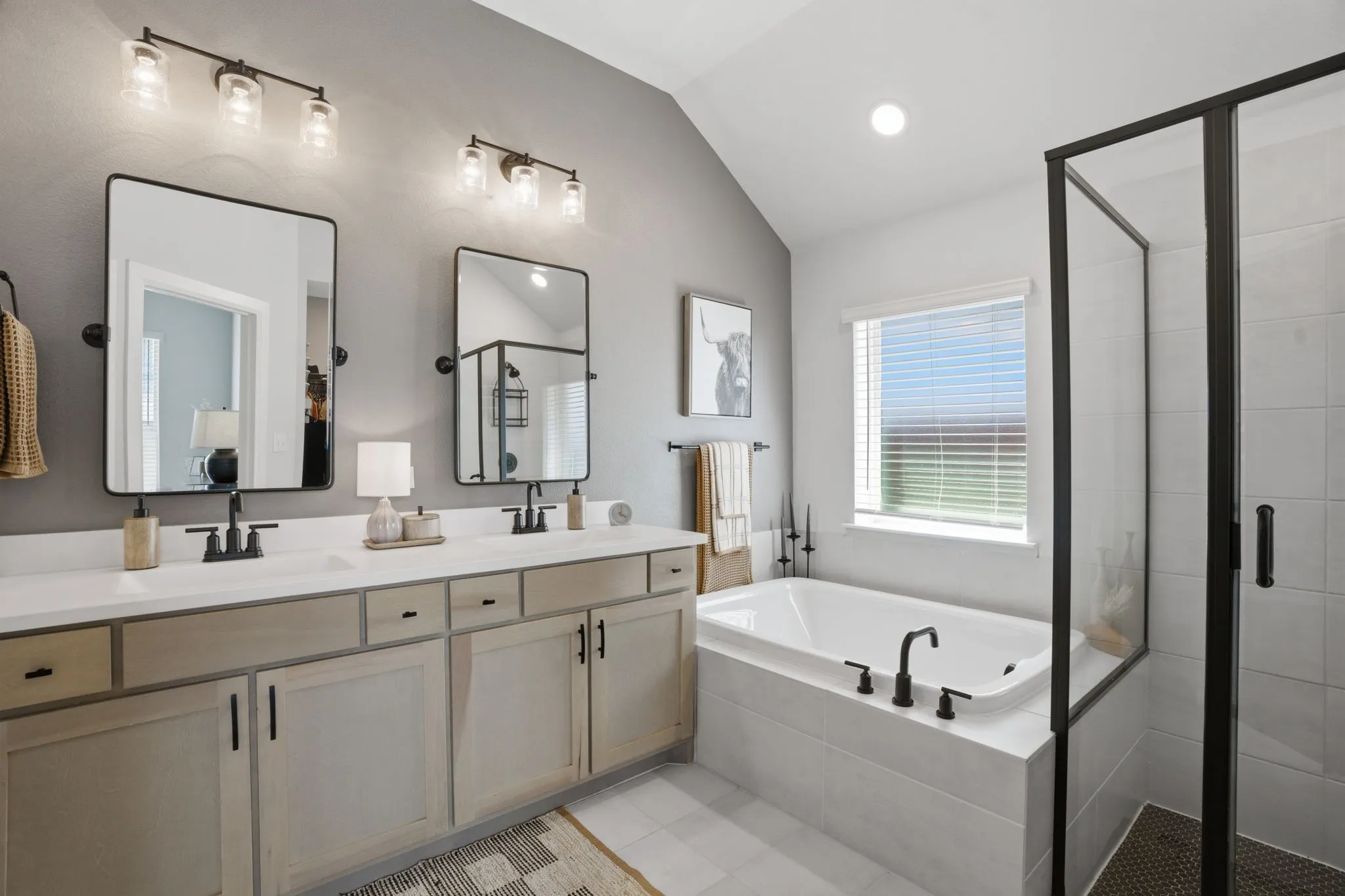 This image showcases a luxurious primary bathroom featuring a spacious design with a double vanity, complete with elegant undermount sinks and a sophisticated aesthetic achieved through the use of sleek black hardware. The cabinetry, finished in a warm, light wood, adds a touch of natural elegance, complementing the overall refined ambiance. The bathroom also boasts a separate, inviting shower area and a relaxing deep garden tub, perfect for unwinding after a long day.