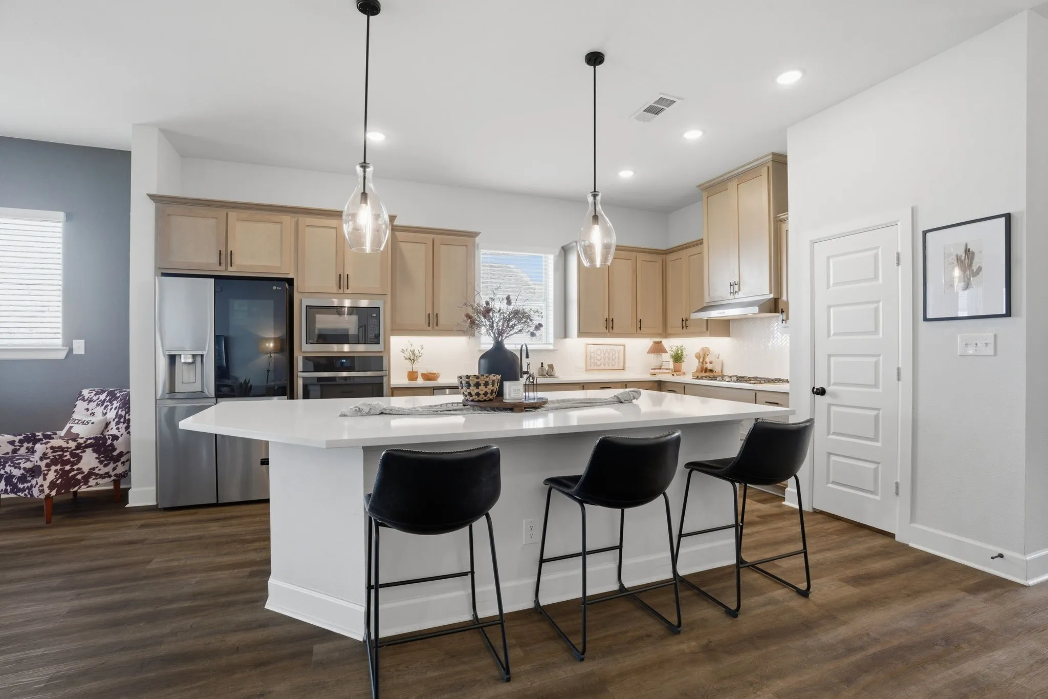 This photo showcases a desirable and contemporary open-concept kitchen, characterized by its spacious design and inviting features. The kitchen boasts a large, expansive island, perfect for both meal preparation and casual gatherings, complemented by a crisp, white countertop that provides a clean and bright aesthetic. A convenient breakfast bar seating area is integrated into the island, encouraging informal dining and social interaction. High-quality stainless steel appliances add a touch of modern elegance and efficiency, while the light wood cabinetry creates a warm and inviting atmosphere, contrasting beautifully with the sleek appliances. The wood-look flooring throughout the space ties the design together, offering both visual warmth and a practical, easy-to-maintain surface.