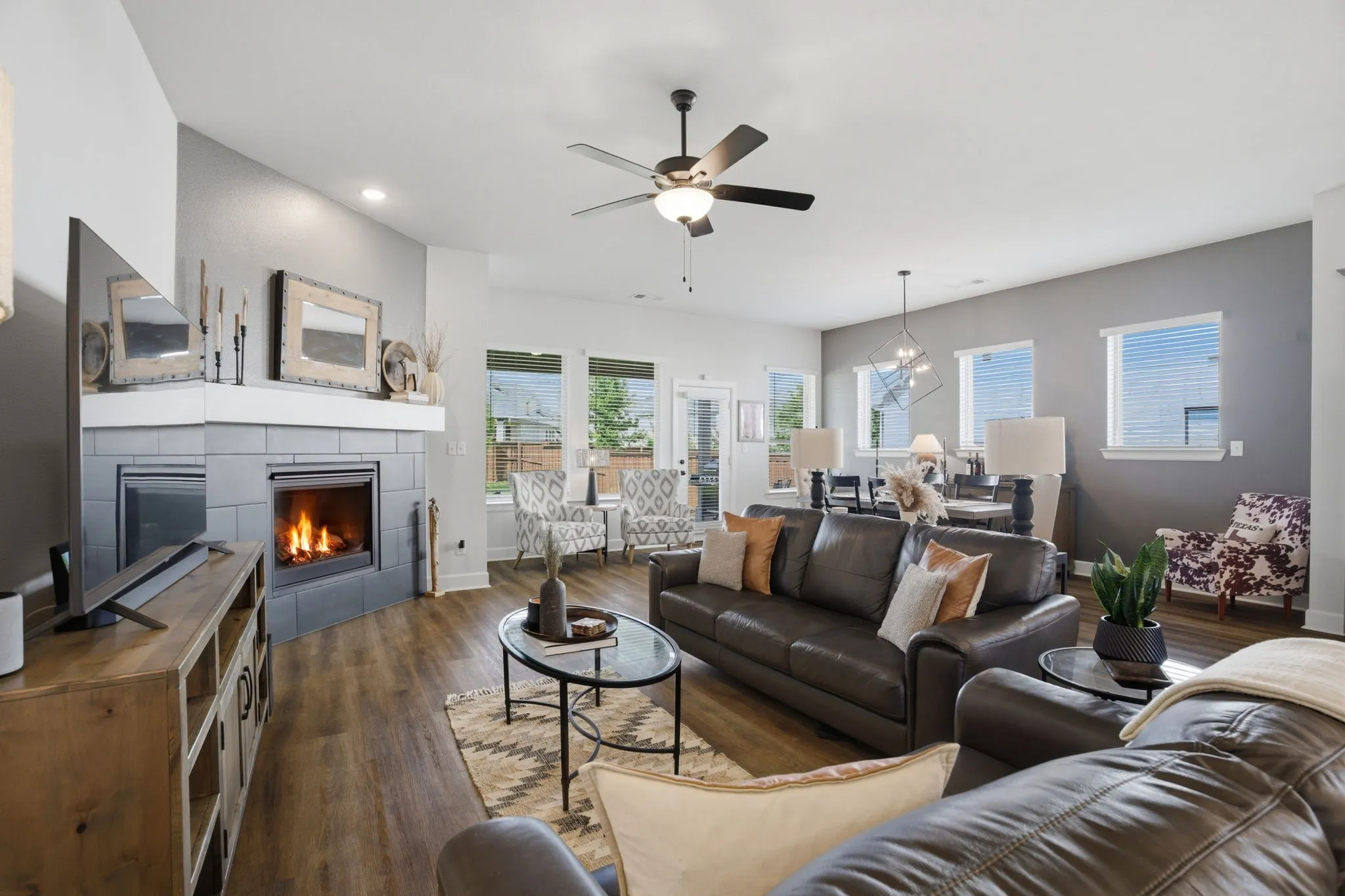 This is a bright and inviting open-concept living area, highlighted by a modern fireplace that boasts a dark-toned tile backsplash complemented by a crisp, white mantel. The space is further enhanced by the strategic placement of recessed lighting, ensuring a warm and even glow, and numerous windows, which flood the room with natural light, creating a truly expansive and comfortable atmosphere.