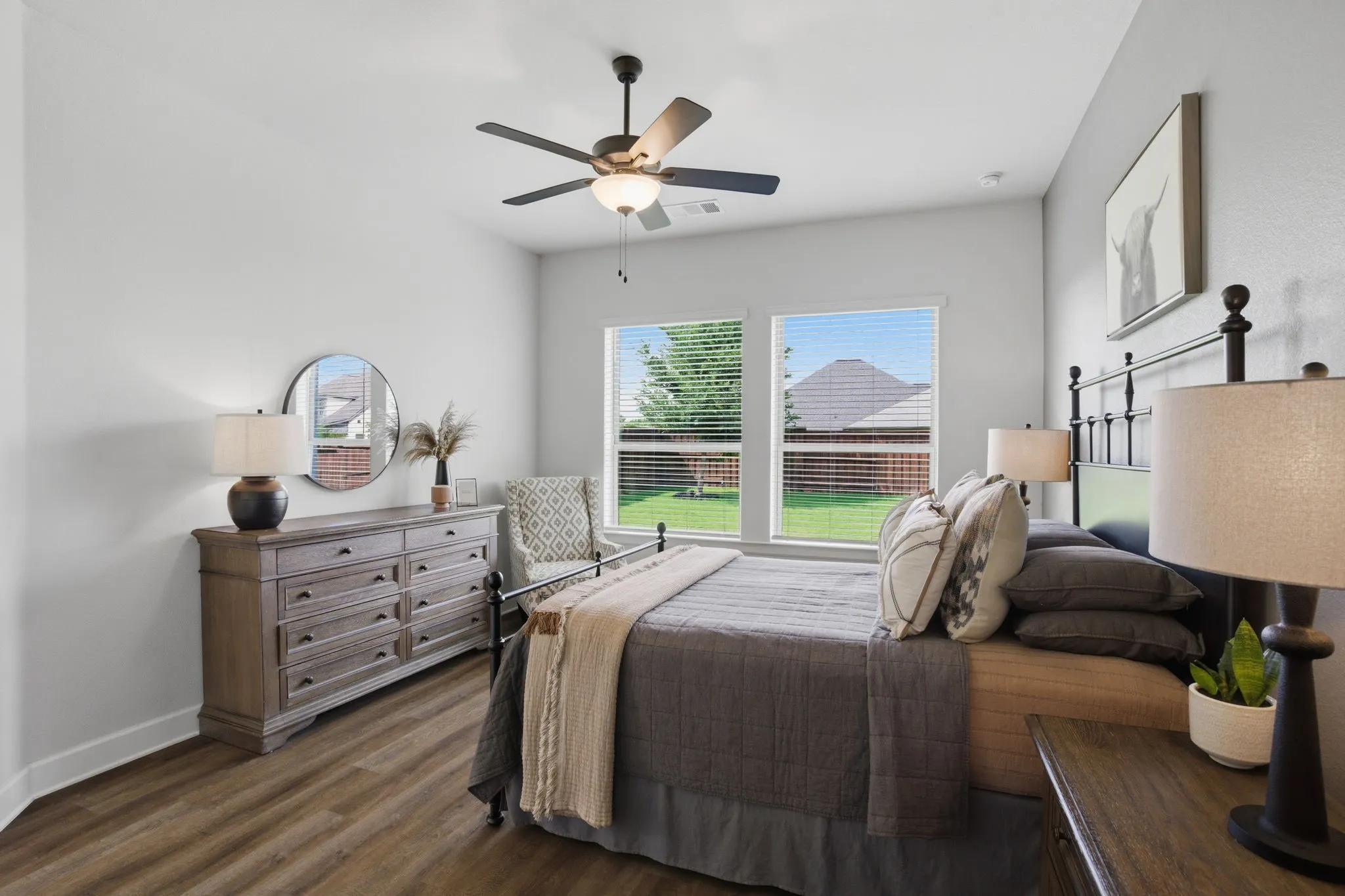 This photo showcases a comfortable and well-designed primary bedroom, highlighting features such as inviting wood-look flooring, soothing light gray walls that create a calming atmosphere, two generously sized windows adorned with practical blinds for light control, a designated and comfortable sitting area perfect for relaxation, and a substantial wall space ideally suited for a large dresser, offering ample storage and visual appeal.