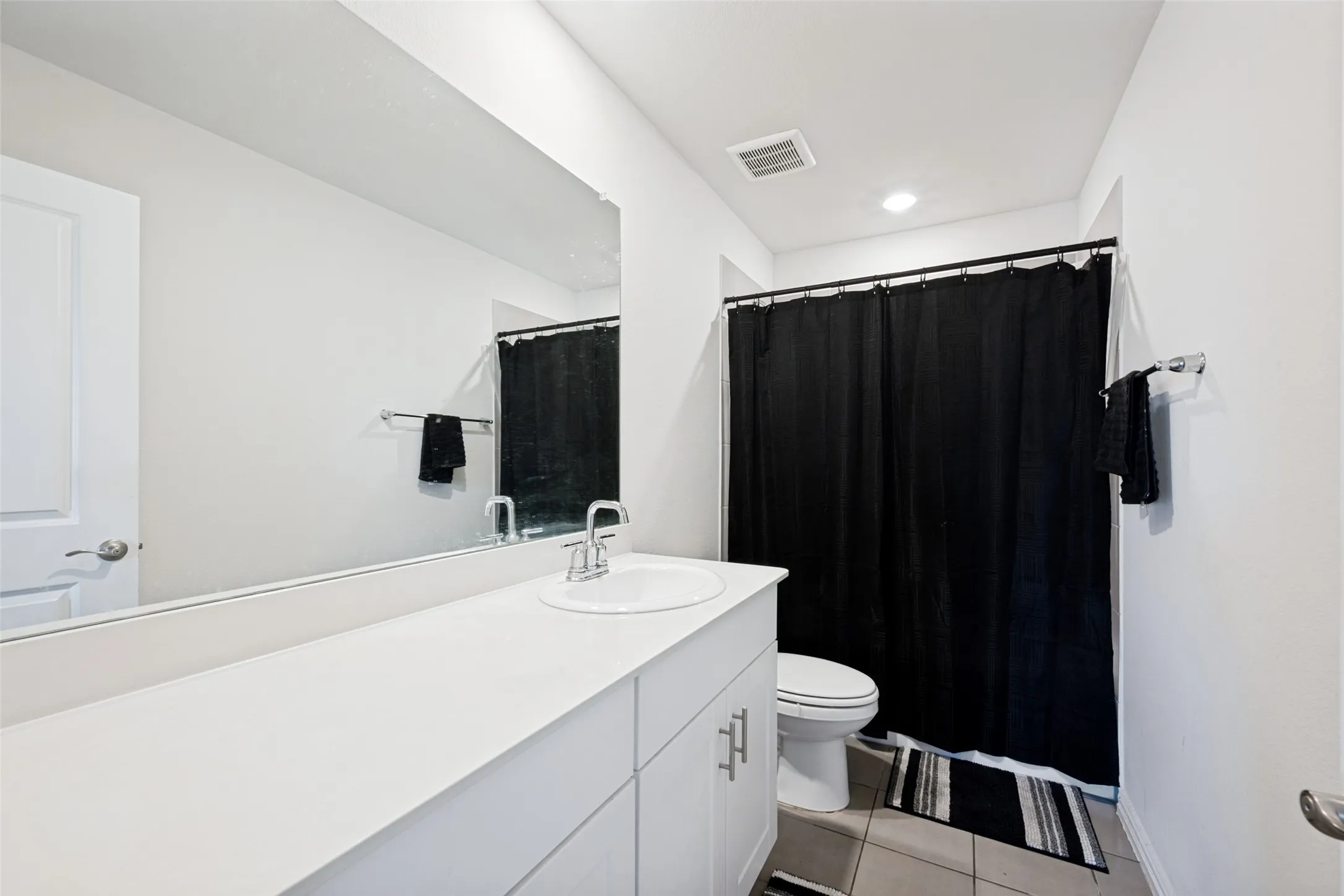 Bathroom featuring a single vanity with an integrated sink, a large wall-mounted mirror, white cabinetry with silver-tone hardware, and a shower-tub combination behind a black curtain