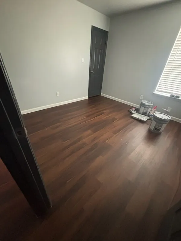 Spacious room featuring wood-finish flooring, light grey walls, white baseboards, and a dark paneled door
