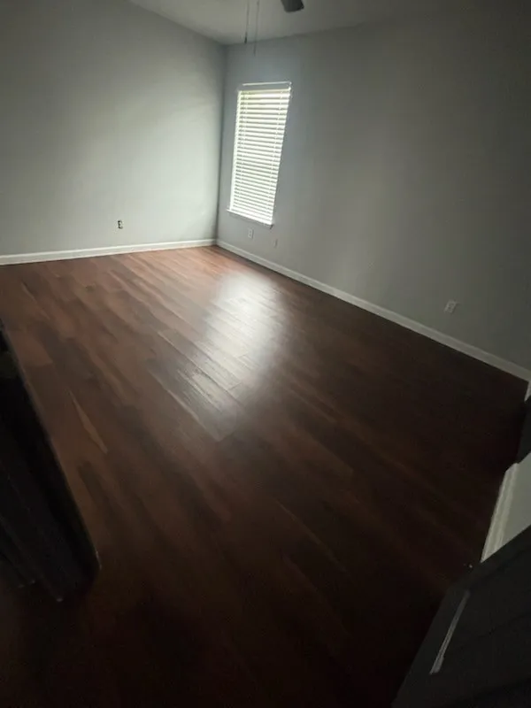 Spacious room featuring rich wood-finish flooring, light gray wall paint, and a single window with horizontal blinds