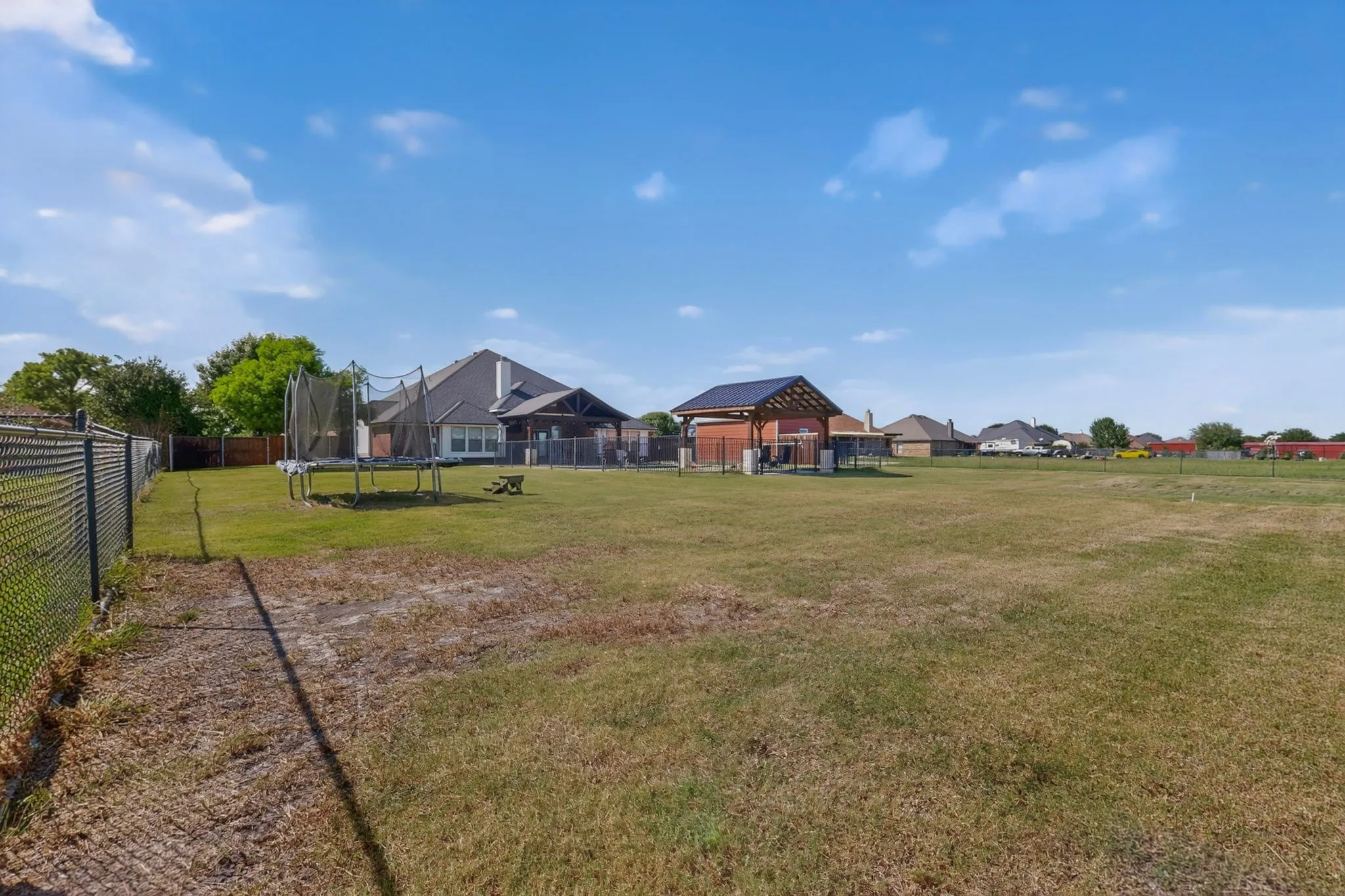 Expansive backyard featuring a large grassy area, chain-link fencing, and a detached open-air pavilion structure with a dark roof