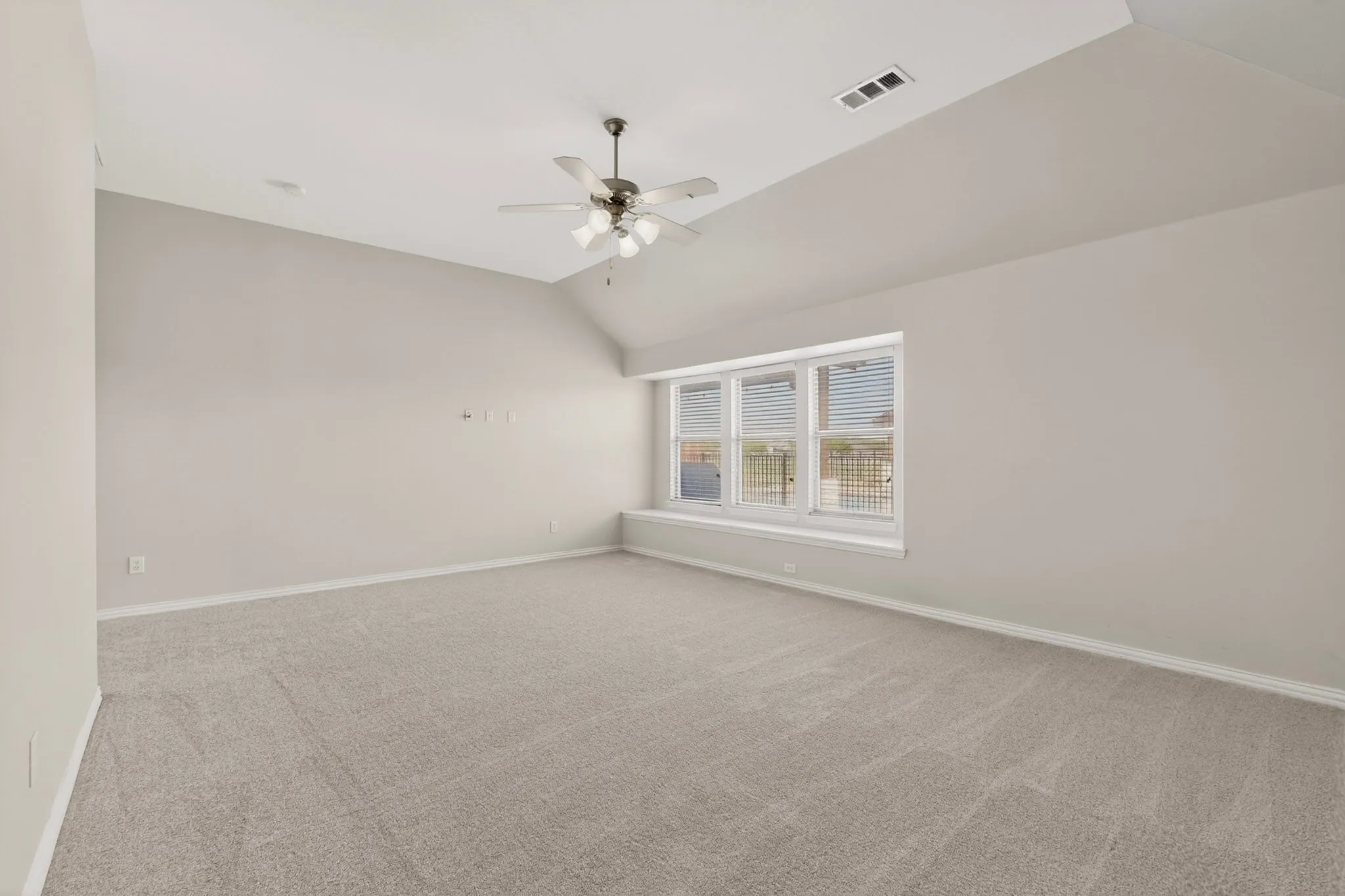 Spacious room featuring vaulted ceilings, light gray carpeting, a large window with blinds, a window seat, and a ceiling fan with integrated lighting