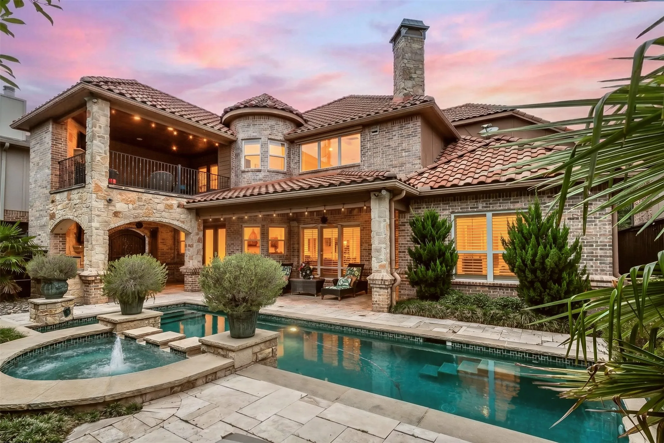 Mature landscaping, privacy fencing, and extensive stone and brick architecture create a private resort atmosphere, while the twilight setting beautifully highlights the home's exceptional outdoor design and architectural presence.