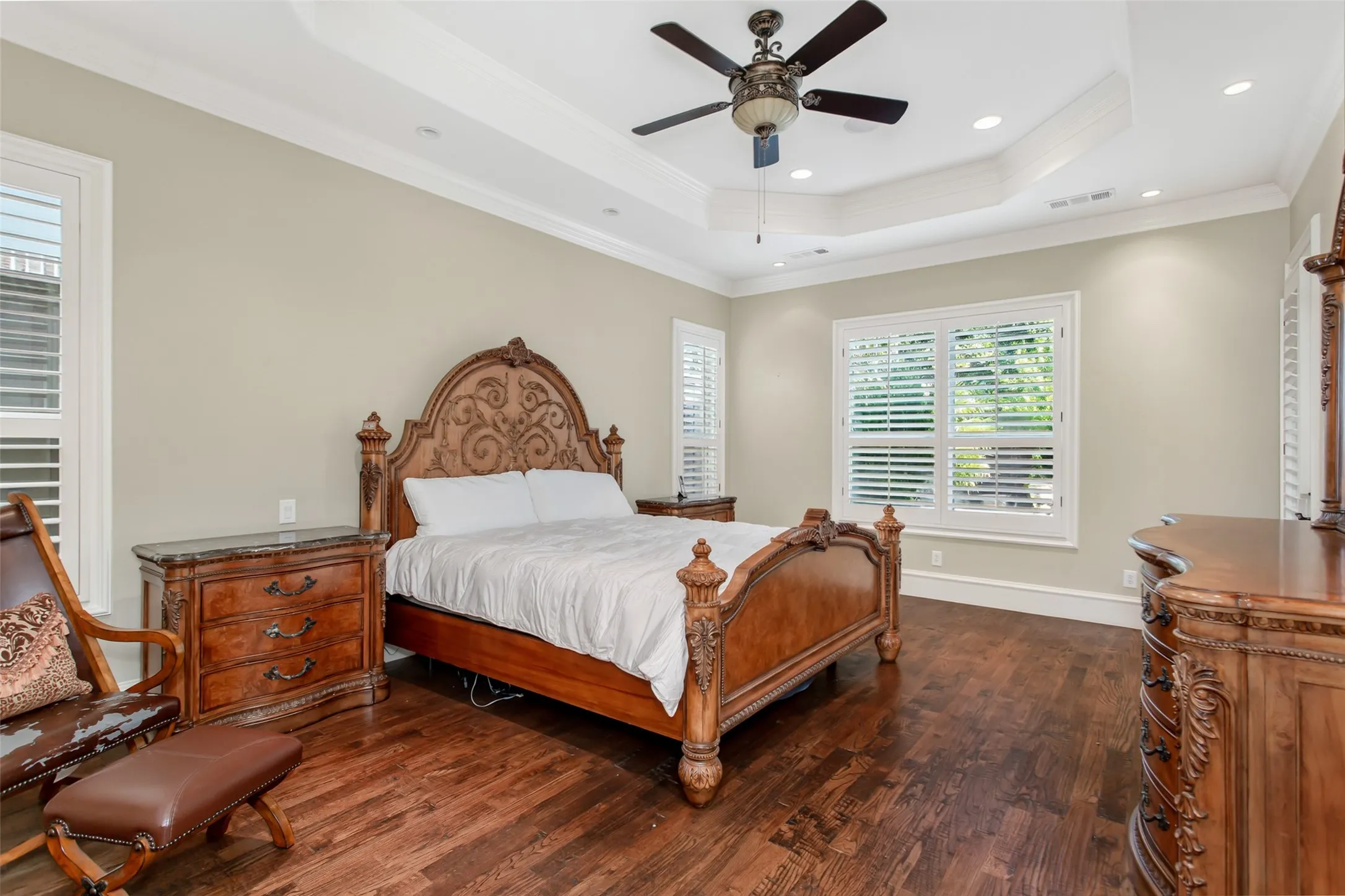 Luxurious first-floor primary suite featuring a dramatic tray ceiling with crown molding, rich hardwood floors, plantation shutters, and generous proportions that easily accommodate oversized furnishings.
