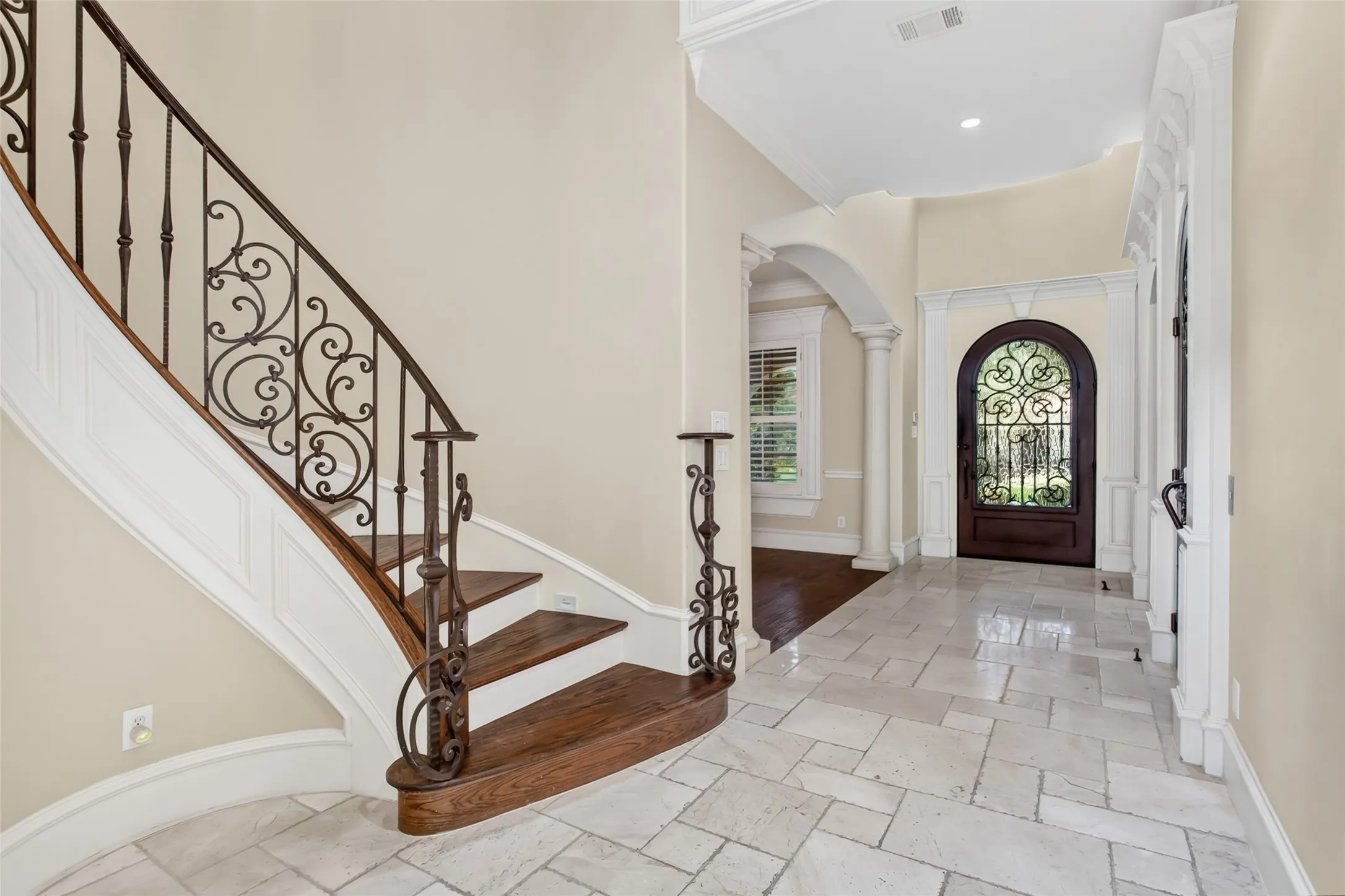 An impressive two-story foyer welcomes guests with travertine flooring, a sweeping staircase with custom wrought-iron balusters, elegant architectural columns, and a handcrafted iron and glass front door that creates a memorable first impression.