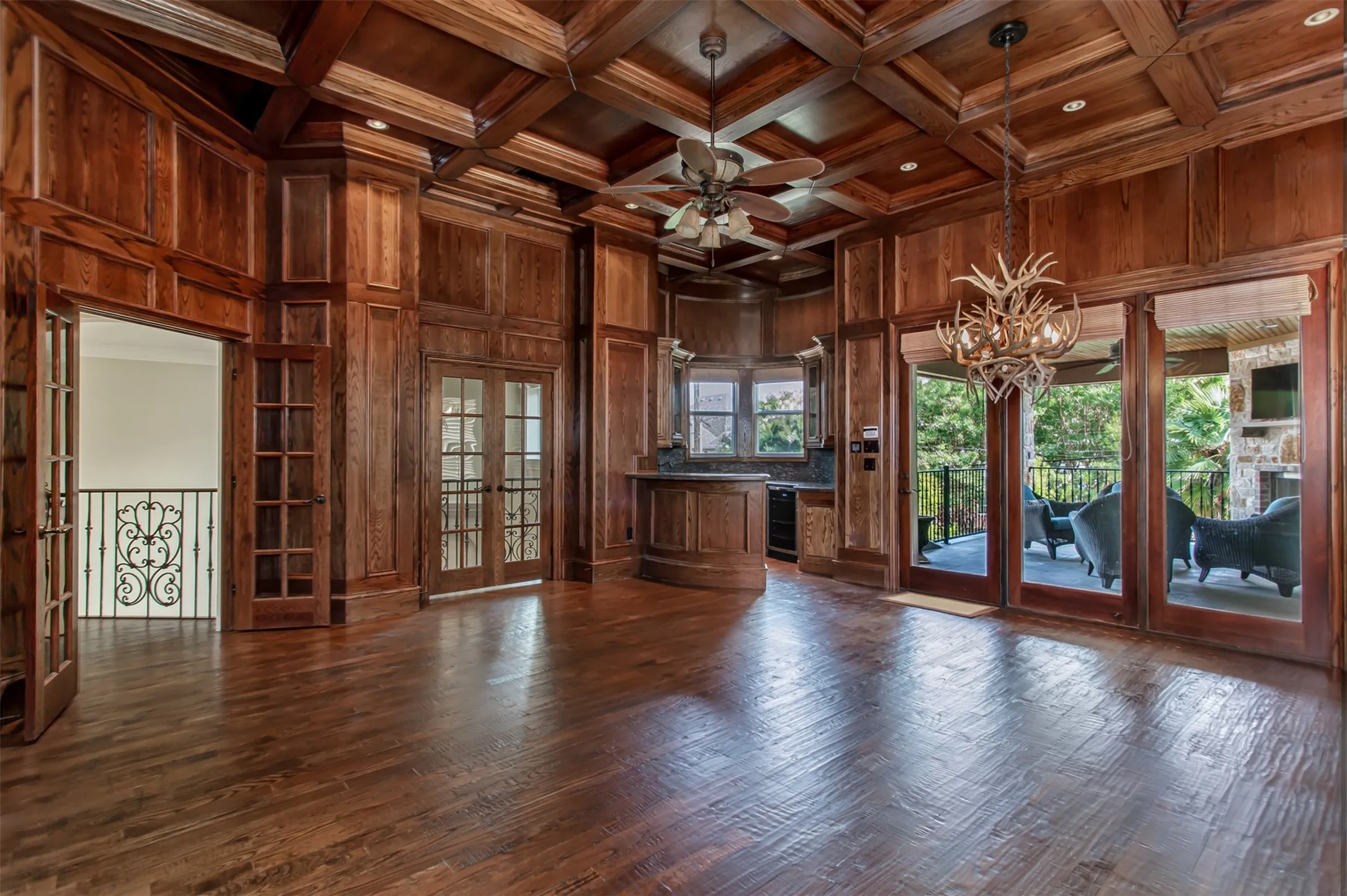 An extraordinary upstairs game room showcases rich floor-to-ceiling wood paneling, a dramatic coffered wood ceiling, and a striking antler chandelier. The space is complete with a built-in wet bar featuring granite countertops, sink, beverage refrigerator, and custom cabinetry, while French doors open to a covered balcony overlooking the resort-style backyard—creating the perfect setting for entertaining.