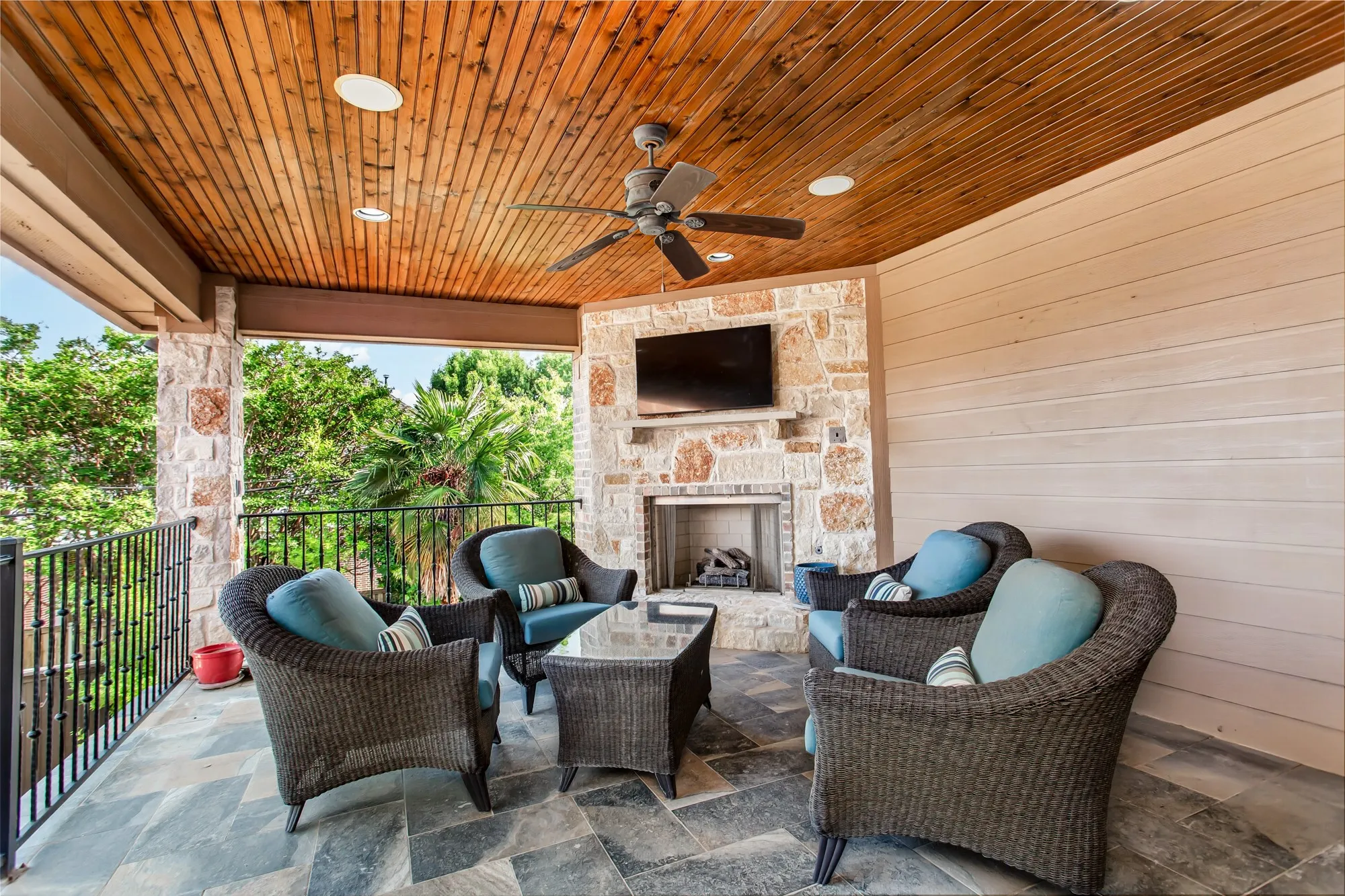 Just off the game room, a spacious covered balcony extends the entertainment space outdoors. The balcony features a wood-plank ceiling with recessed lighting, ceiling fan, built-in speakers, and a stone fireplace with mounted television, creating an inviting setting for year-round relaxation. From this elevated vantage point, enjoy sweeping views of the resort-style backyard, including the custom pool and raised spa with multiple water features, expansive travertine decking, and lush landscaping below.
