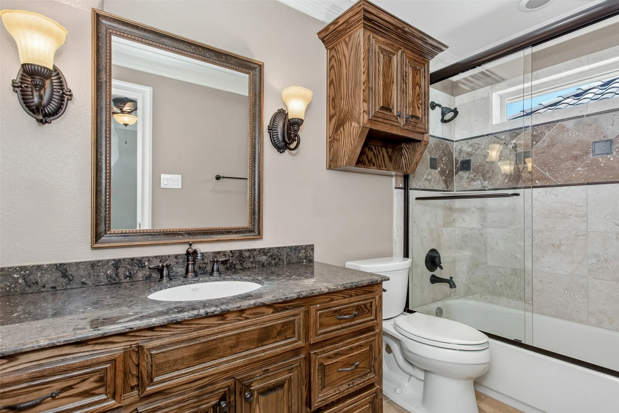 The private bath features a granite-topped vanity, custom wood cabinetry, designer lighting, and a tub/shower combination with beautifully tiled surround and glass enclosure.