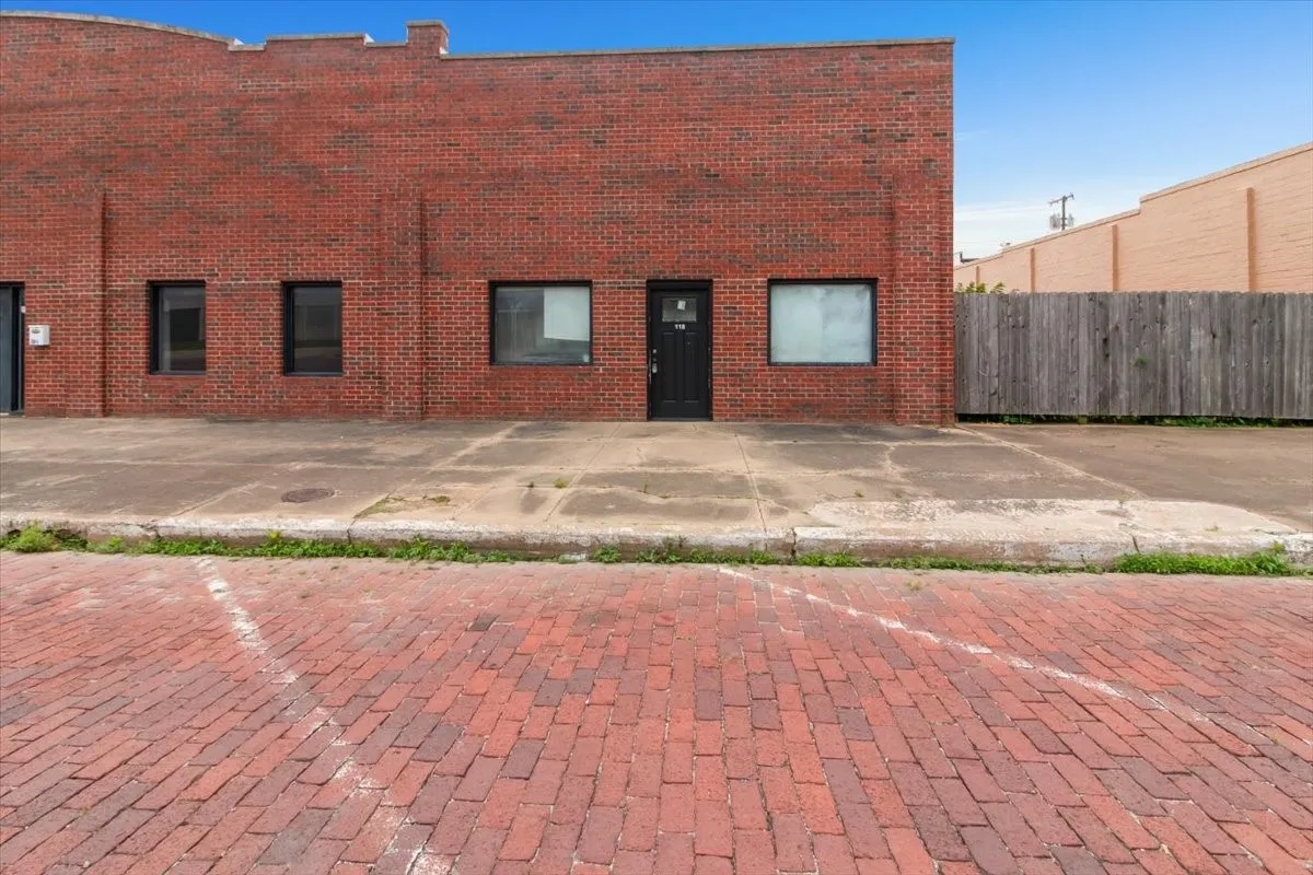 Red brick exterior building featuring multiple window openings, a black entry door, and a concrete sidewalk