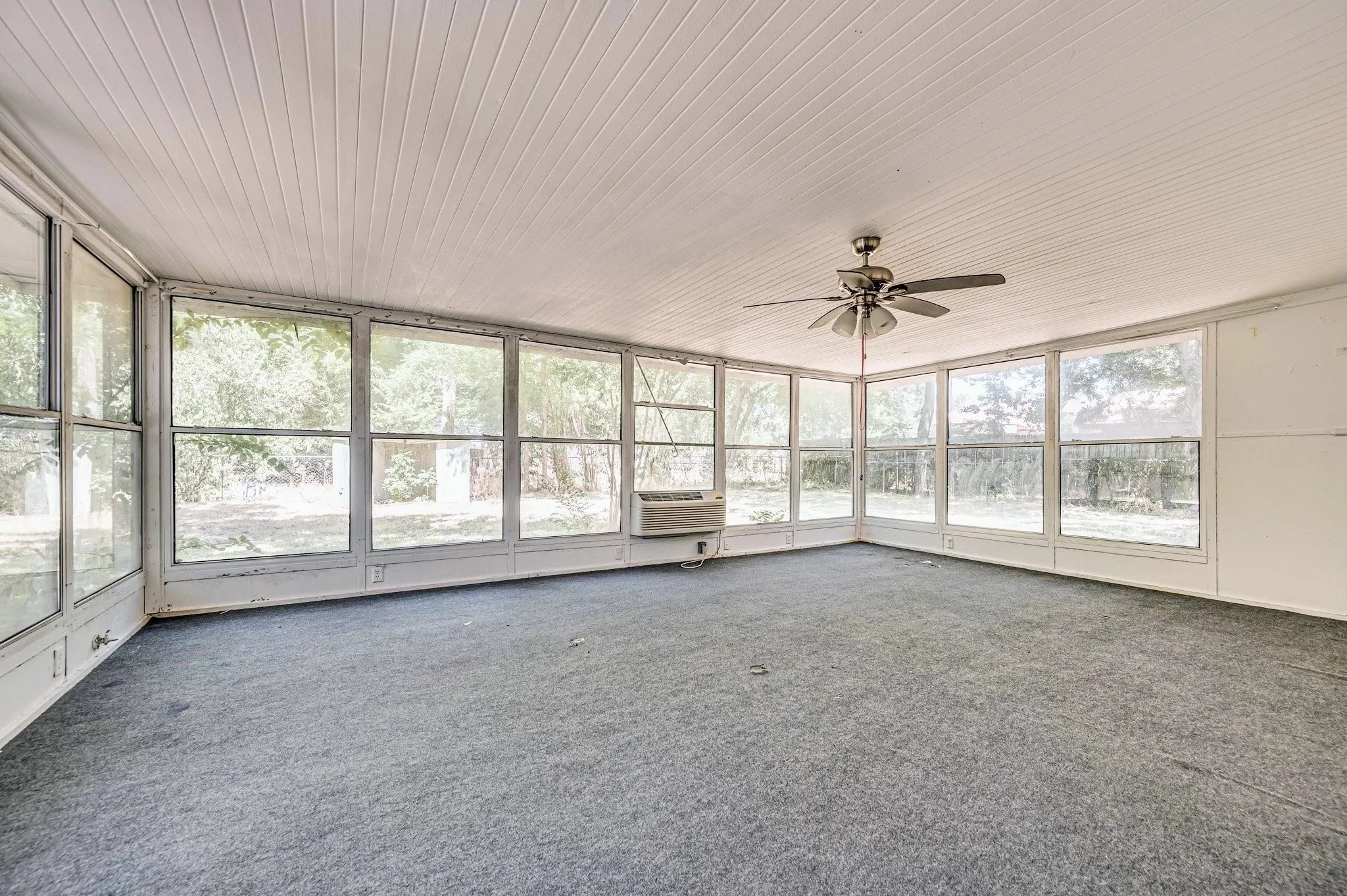 Spacious sunroom featuring a paneled ceiling, expansive windows, wall-mounted air conditioning unit, ceiling fan, and carpeted flooring