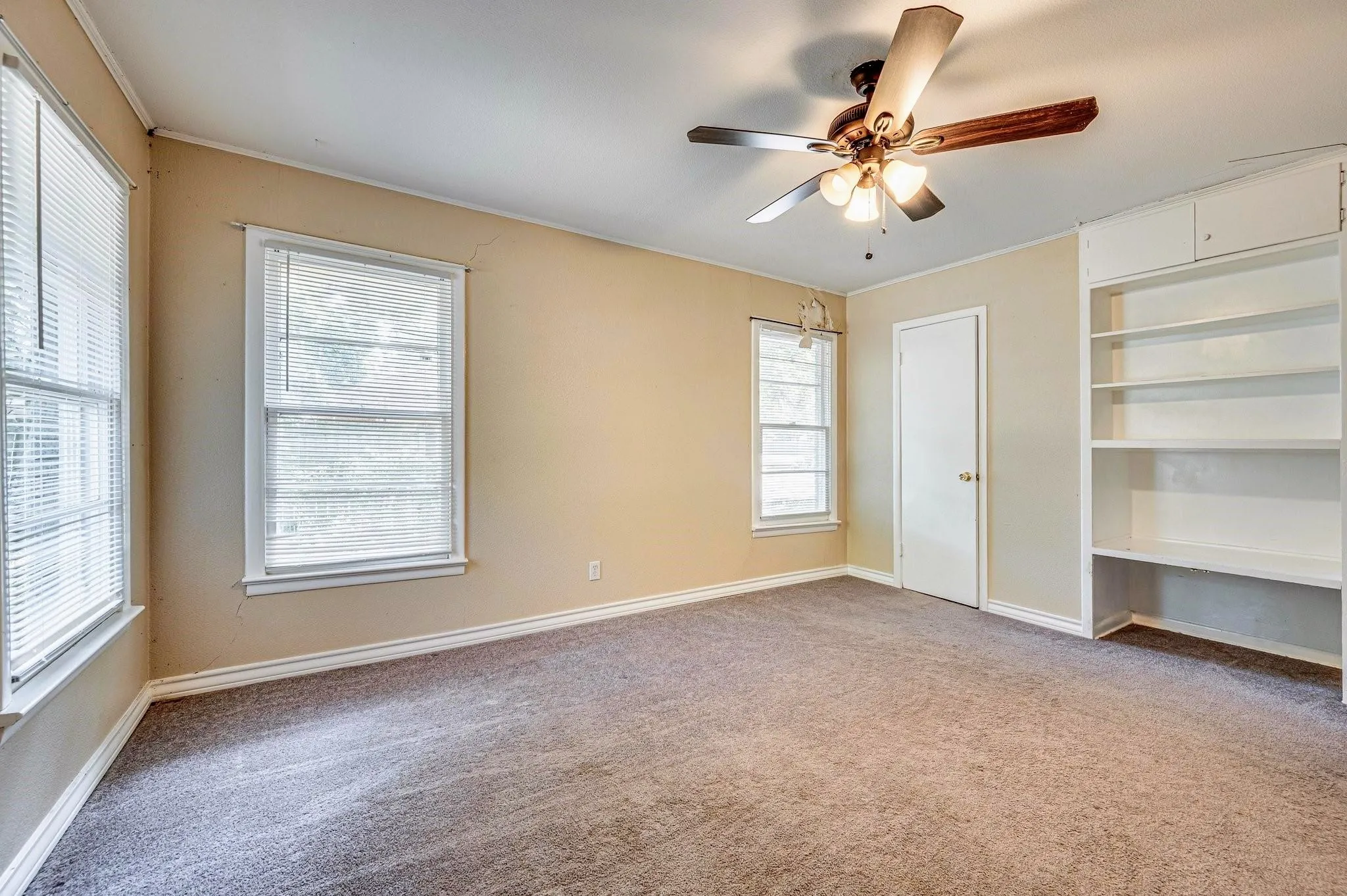 Carpeted room featuring multiple windows with blinds, a ceiling fan with light fixture, built-in shelving, and a white interior door