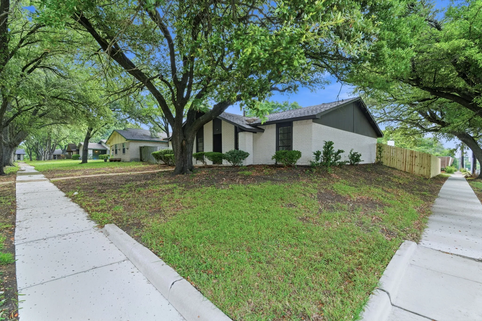 Exterior featuring white brick siding with a dark gabled roofline, mature trees, a freshly installed wood privacy fence, and concrete sidewalks