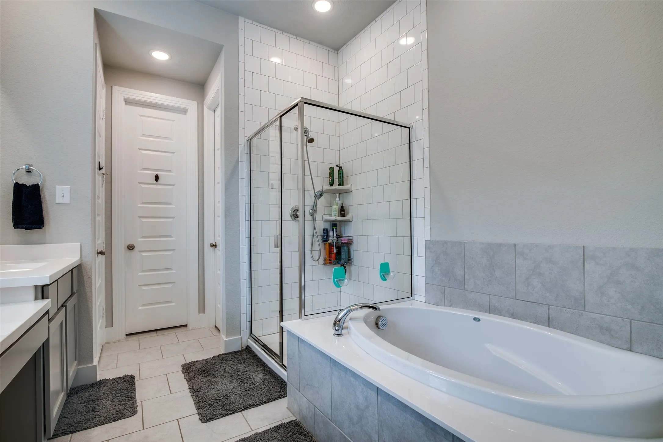 Soaking tub with tile surround, glass-enclosed shower featuring subway tile, recessed lighting, and a dual vanity with white countertops